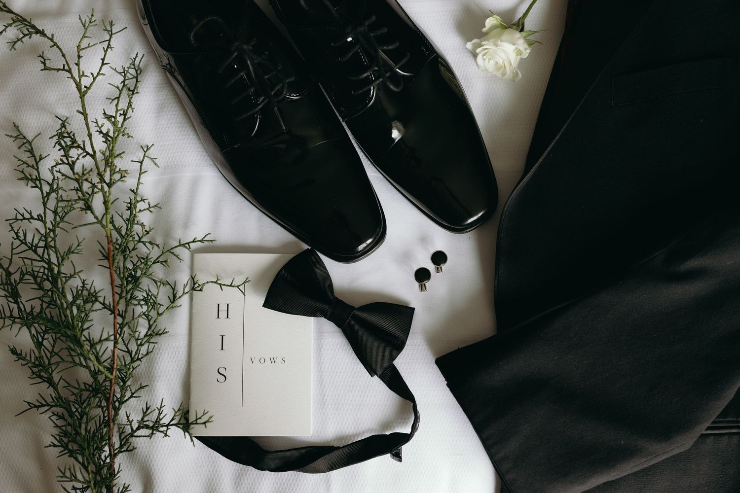 Wedding accessories including black dress shoes, bow tie, and floral decor arranged on white surface.