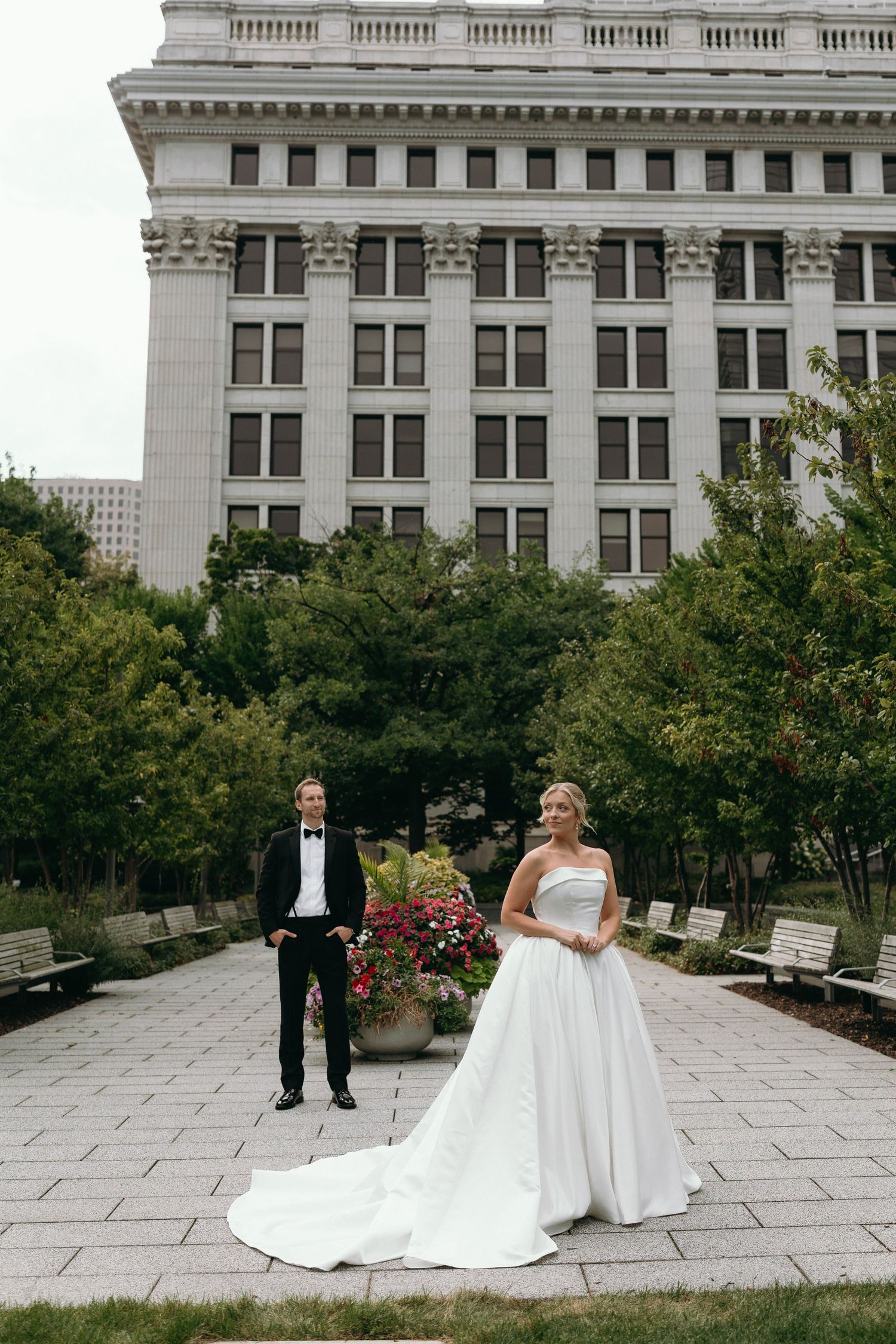 A wedding couple poses on a stone patio with a grand historic building facade in the background.