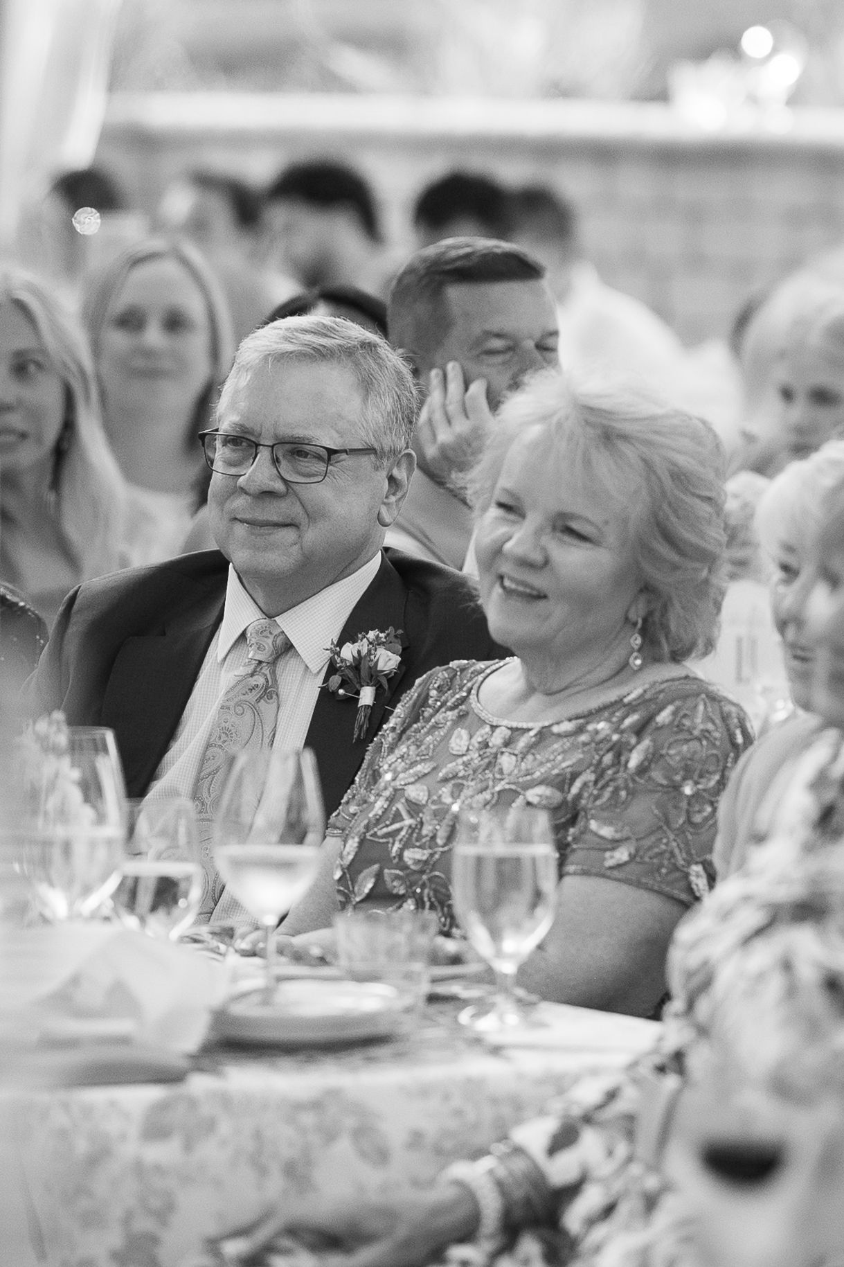 Black and white photo of a smiling older couple at a wedding reception, seated at a decorated table with wine glasses.