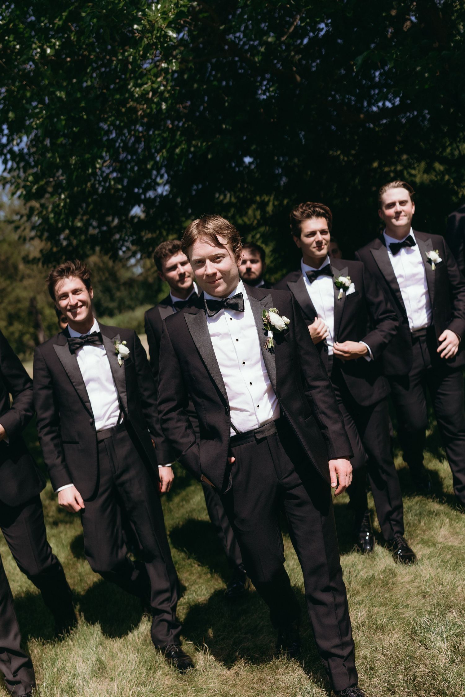 Groomsmen in black tuxedos with bow ties and boutonnieres walk outdoors on a sunny day at a wedding.