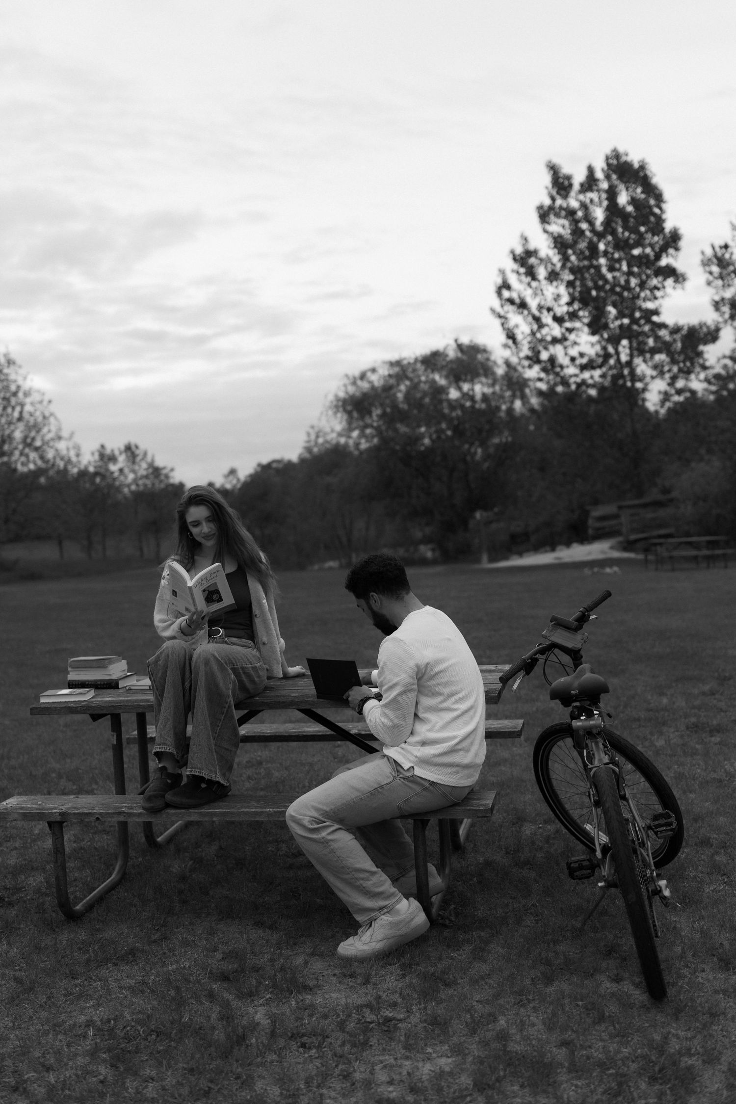 Black and white photo of a couple reading at an outdoor picnic table in a park with a bicycle nearby.