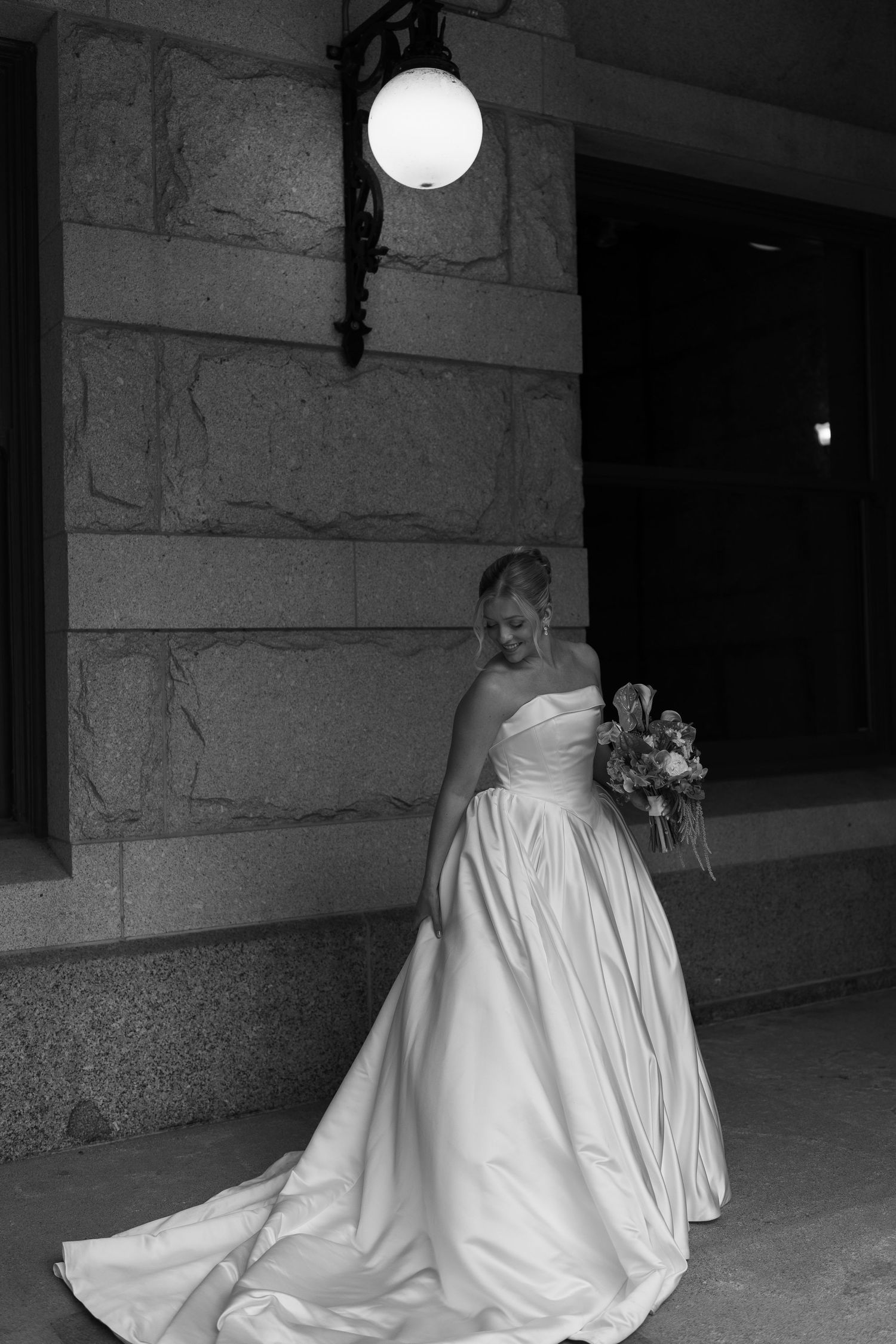 A person in a flowing white wedding gown stands beneath a glowing round light fixture against a stone wall.