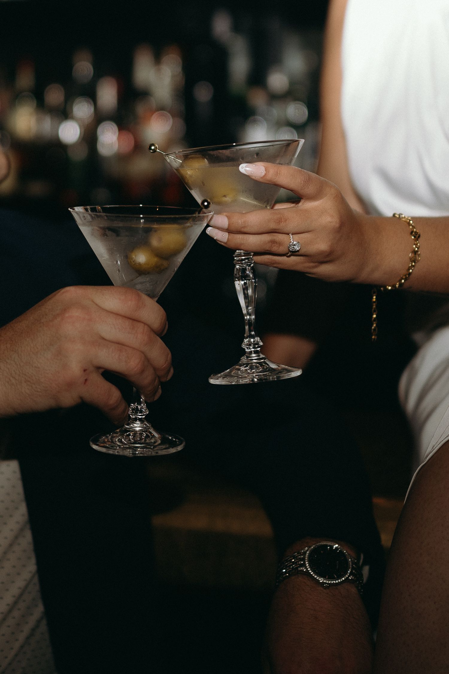 Two martini glasses clink together in dimly lit bar setting.