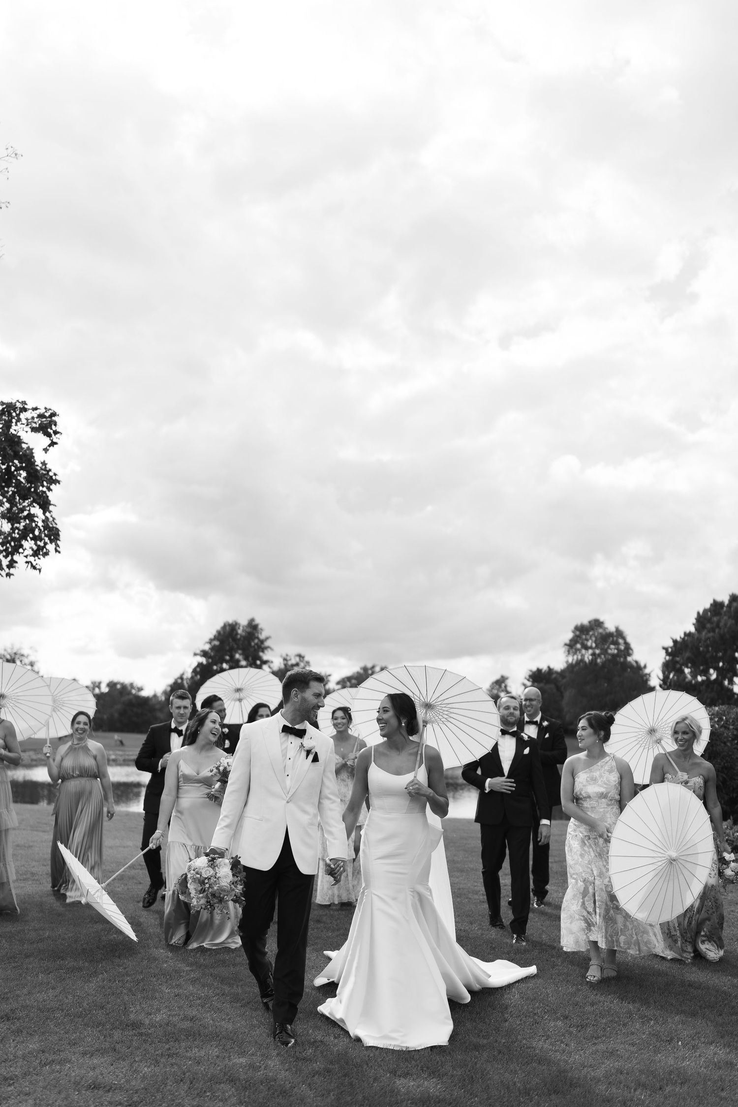 Wedding party walks across lawn with white parasols under cloudy sky.