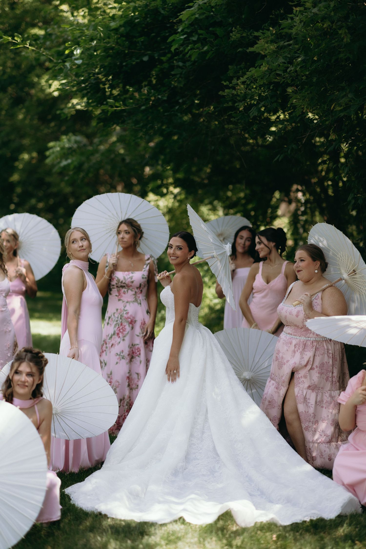 Bride in white gown poses with bridesmaids in pink dresses holding white parasols outdoors.