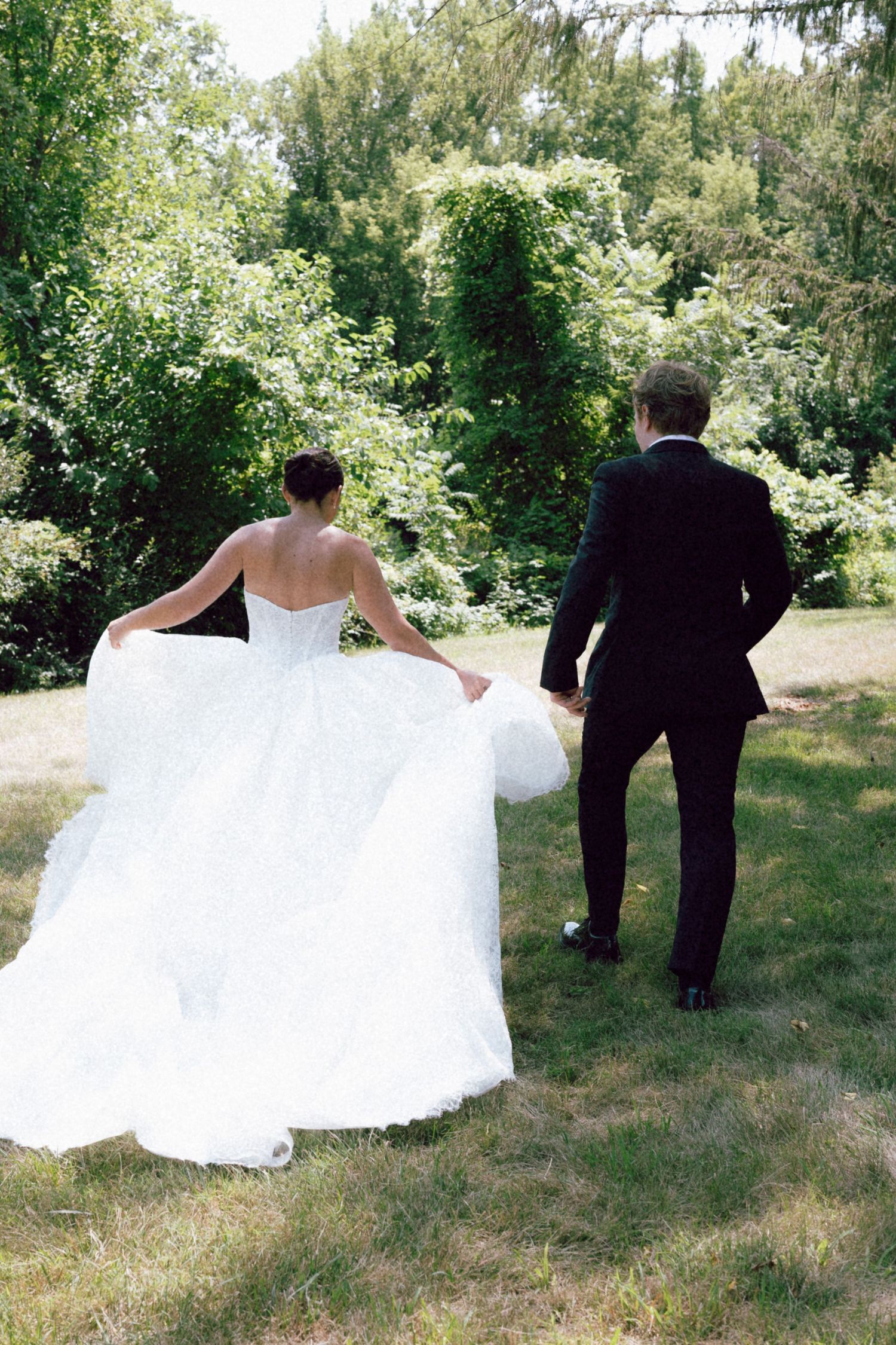 Bride in white gown and groom in black tuxedo walk hand in hand toward lush green trees on wedding day on film