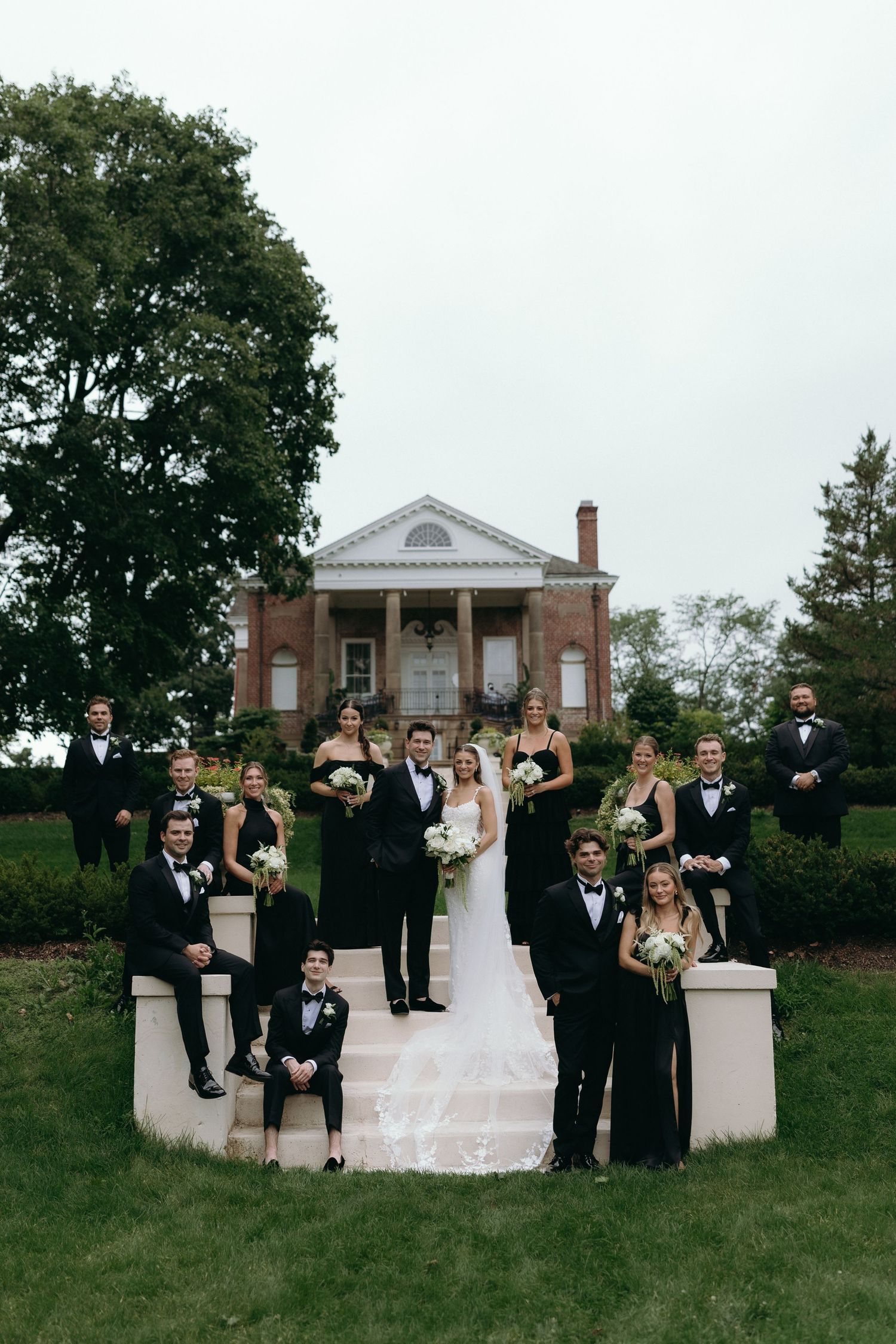 Wedding party in black attire poses on steps before a brick mansion, bride in white gown holding white floral bouquet.