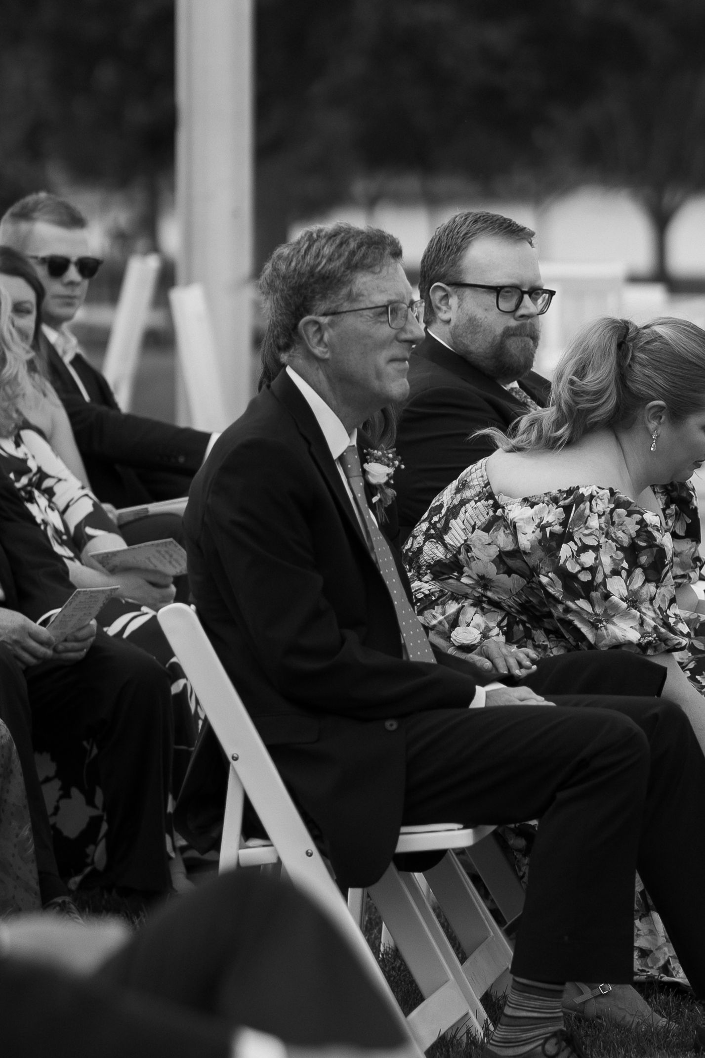 Black and white photo of wedding guests seated in white chairs at an outdoor ceremony.