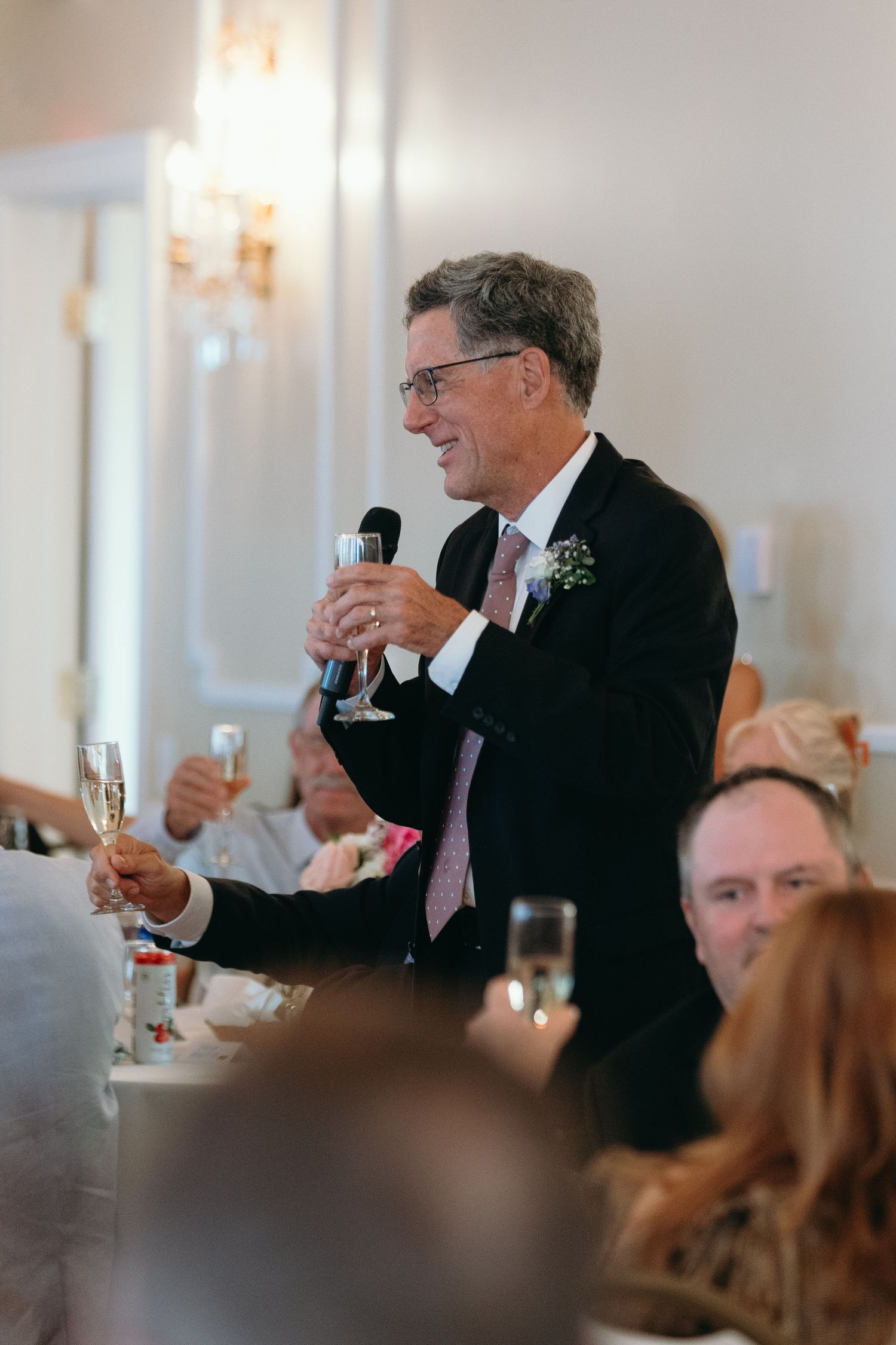 A speaker wearing a dark suit and tie gives a toast with a microphone at an elegant reception with chandeliers.