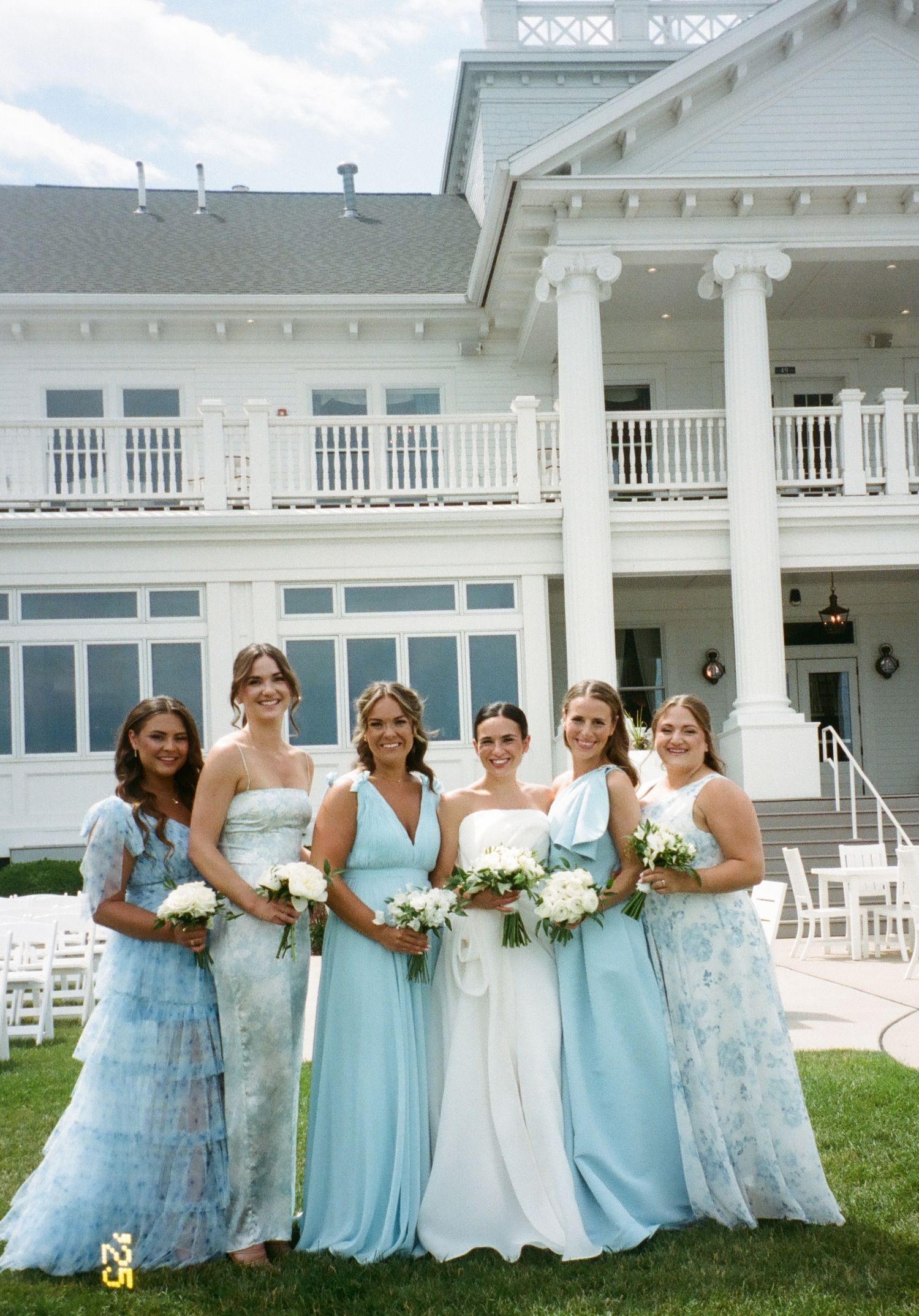 Bridesmaids in light blue dresses pose together on the lawn outside a white wedding venue building.