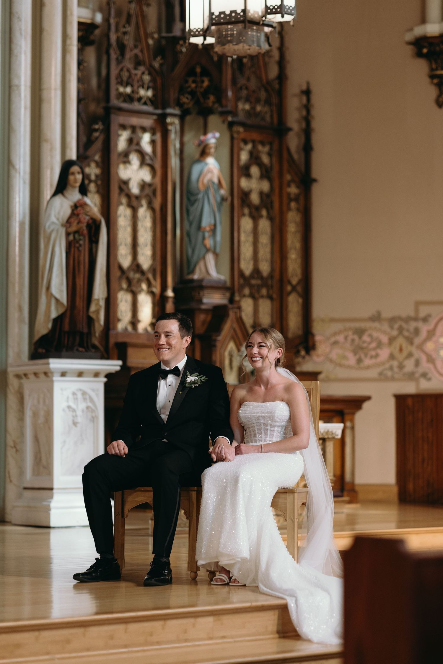 A couple seated at the altar during their wedding ceremony with ornate religious decor behind them.