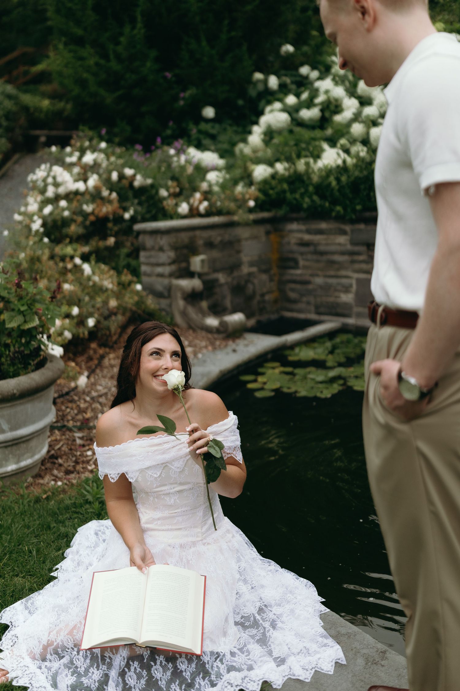 Garden scene with person in white lace dress holding flower by ornamental pond.