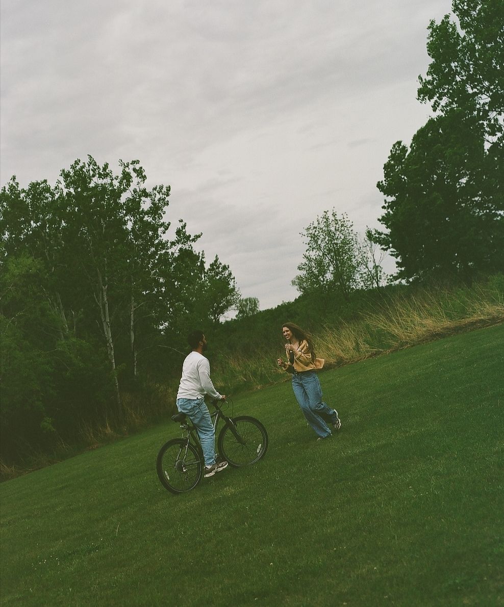 Two people enjoying a green park, one riding a mountain bike on a lush lawn with trees in the background.