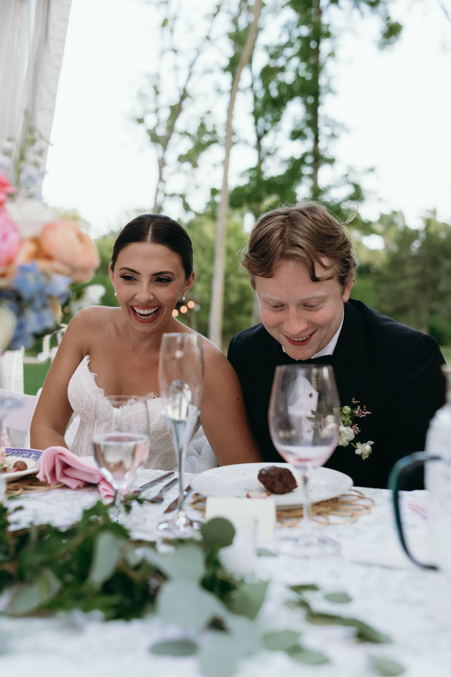 Bride and groom laughing together at wedding reception dinner table with wine glasses.