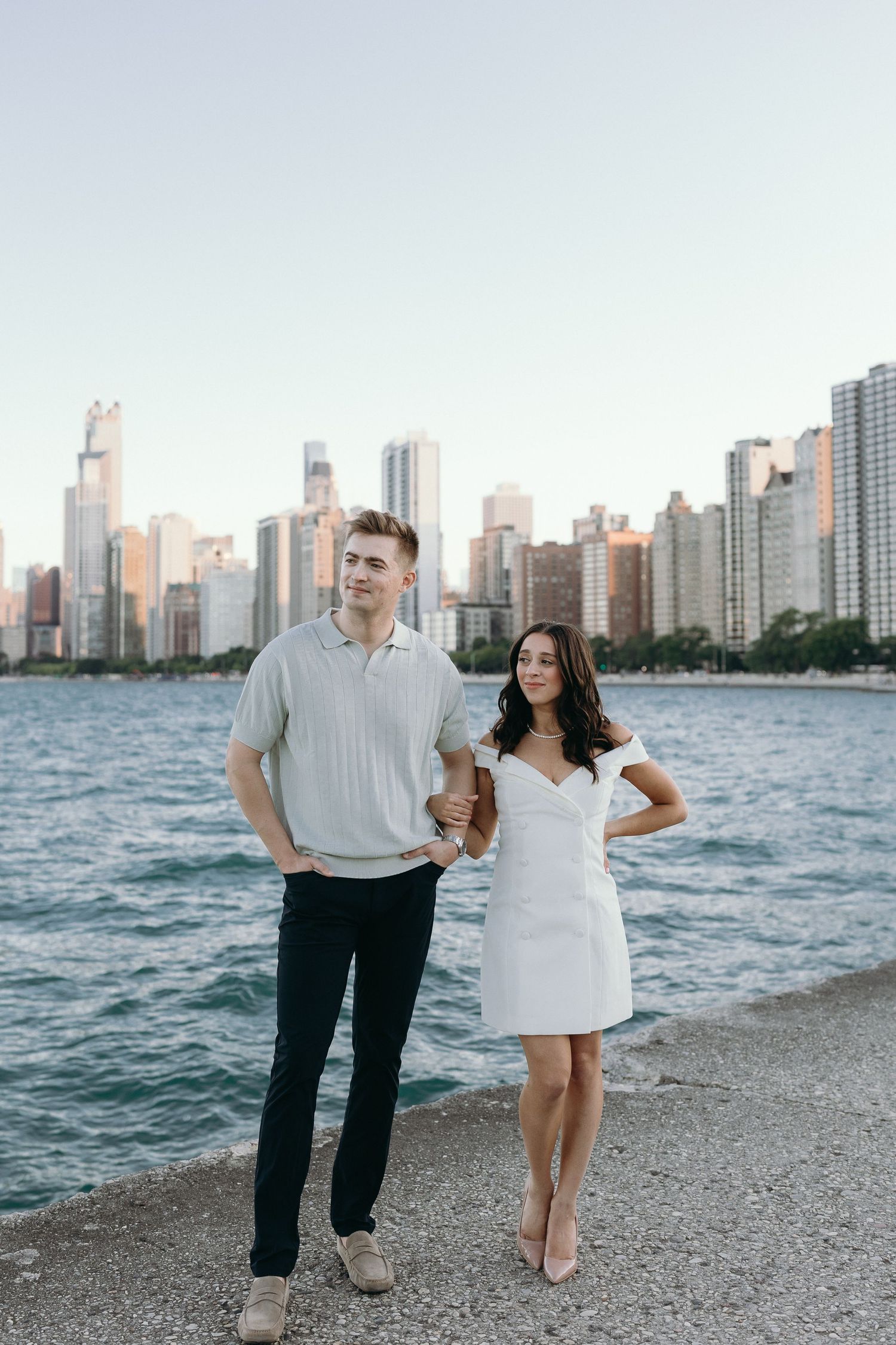 A couple walks along the beach with the Chicago skyline in the background during an engagement photo session.