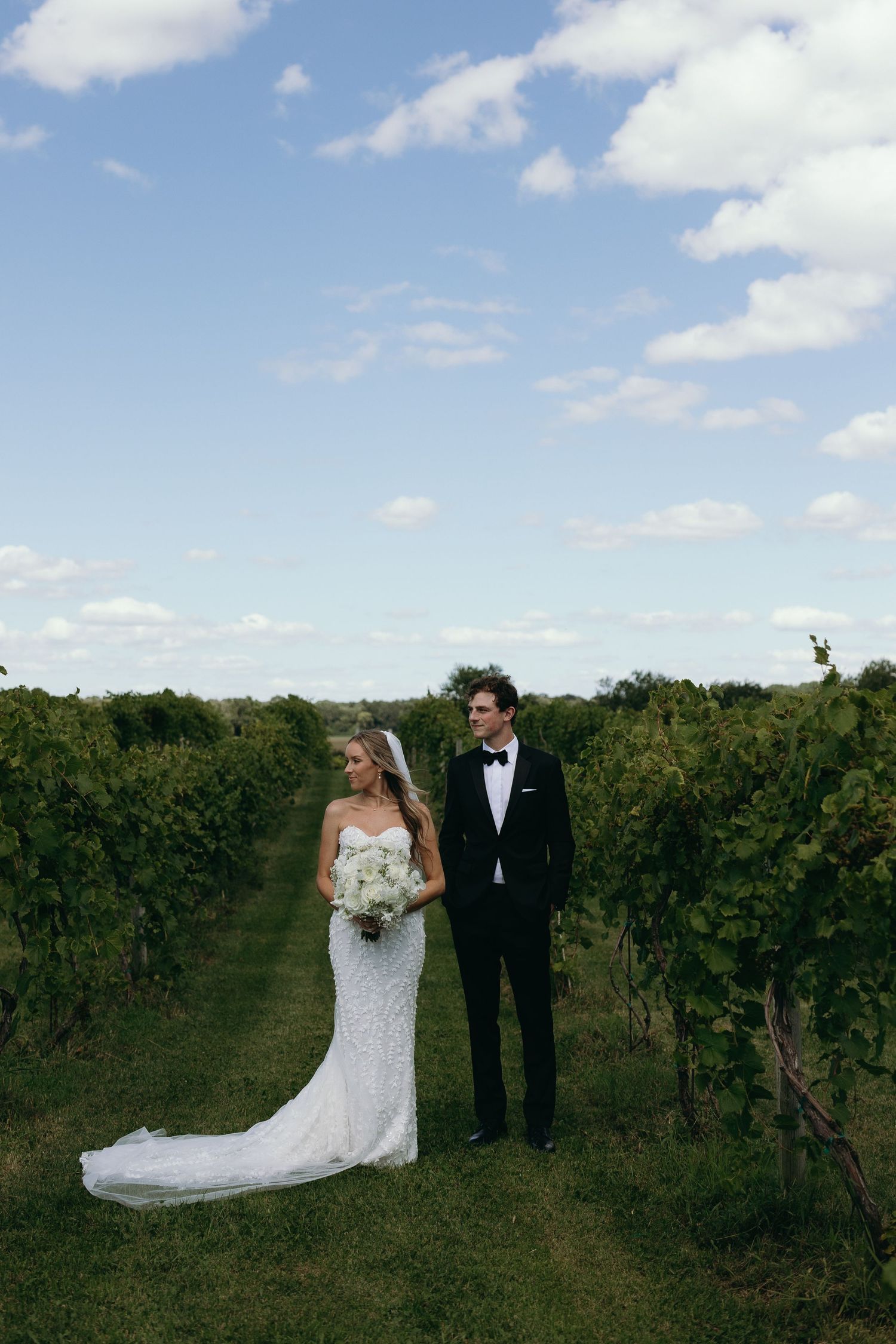 A bride and groom pose together in a lush vineyard on their wedding day under a bright blue sky.
