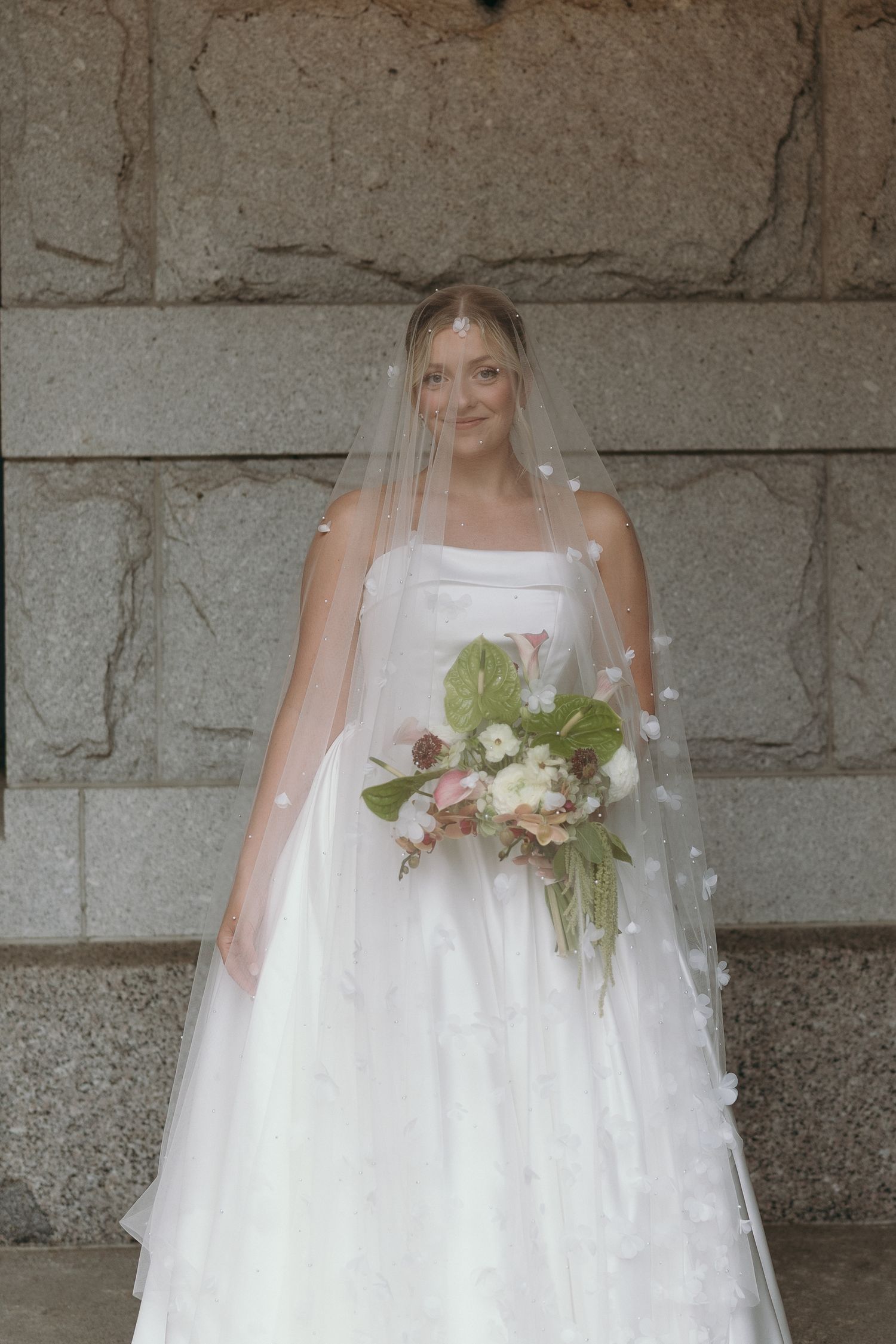 A bride in a white ball gown wedding dress stands against a stone wall holding a white floral bouquet.