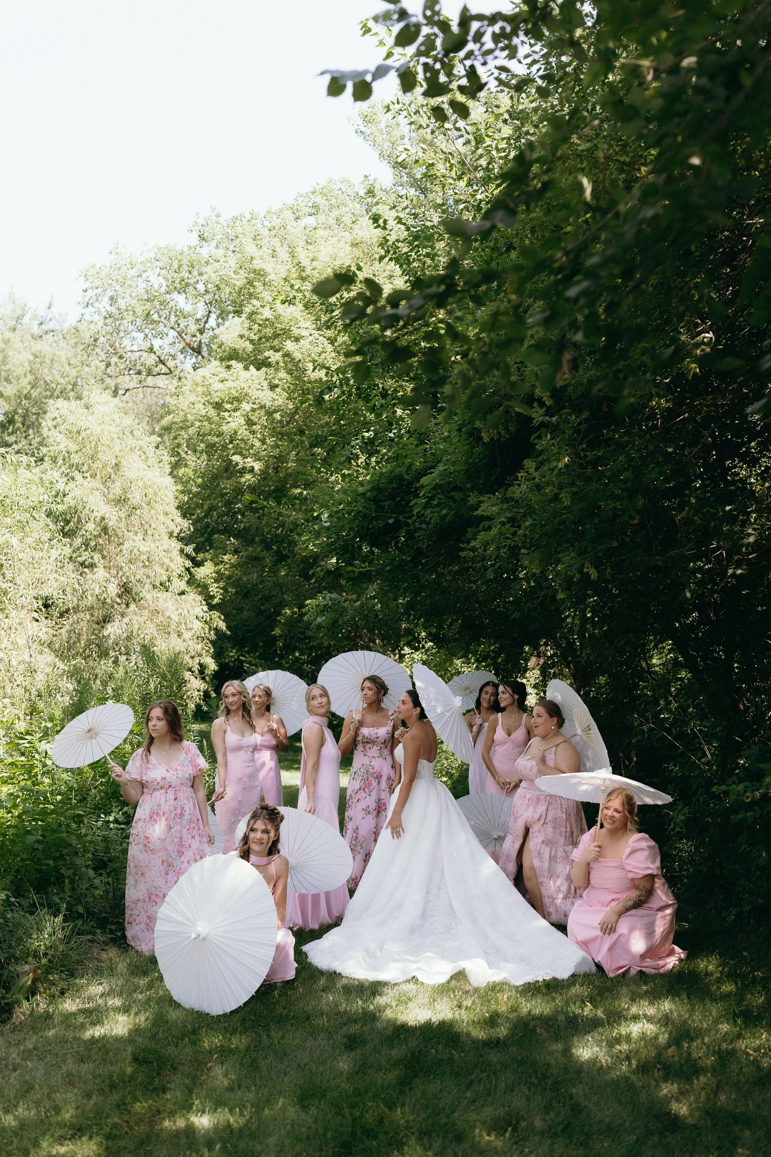 Bride in white gown poses with bridesmaids in pink floral dresses holding white parasols outdoors among lush green trees.