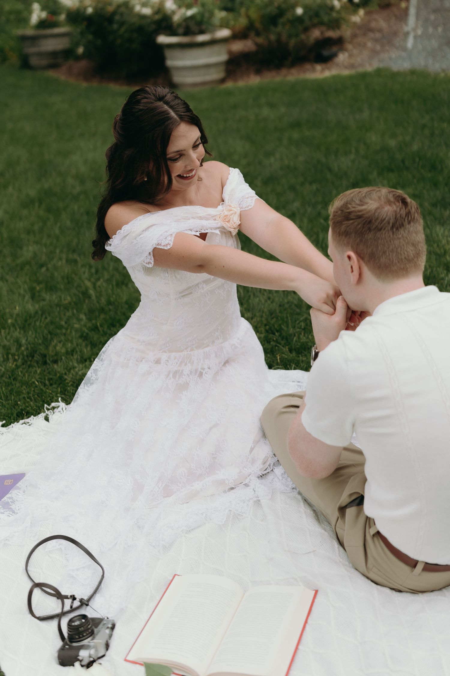A couple in wedding attire sits on picnic blanket playing a game of chess outdoors on a lawn.