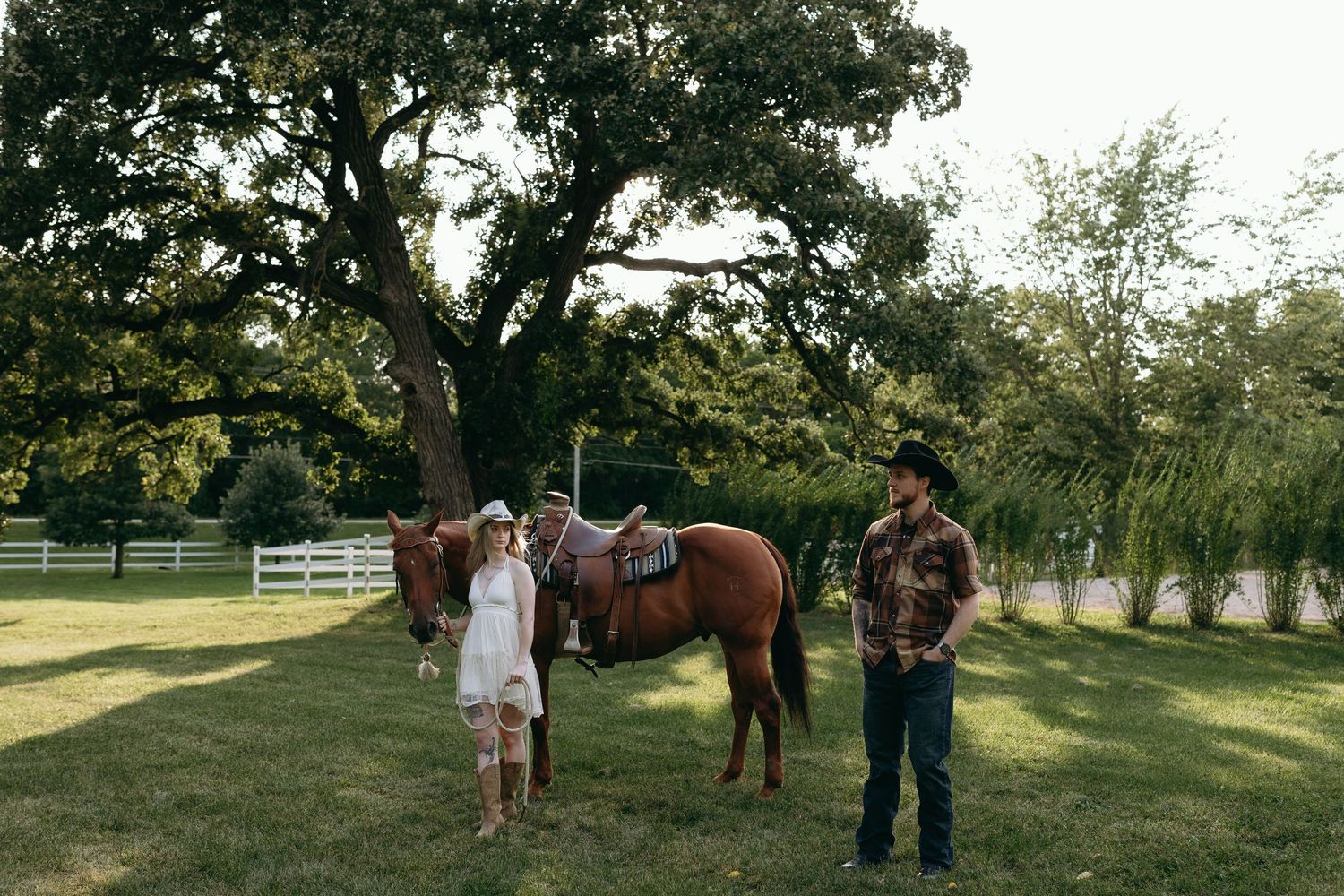 Group gathers outdoors around horses and riders on a sunny day with white fencing in the background.