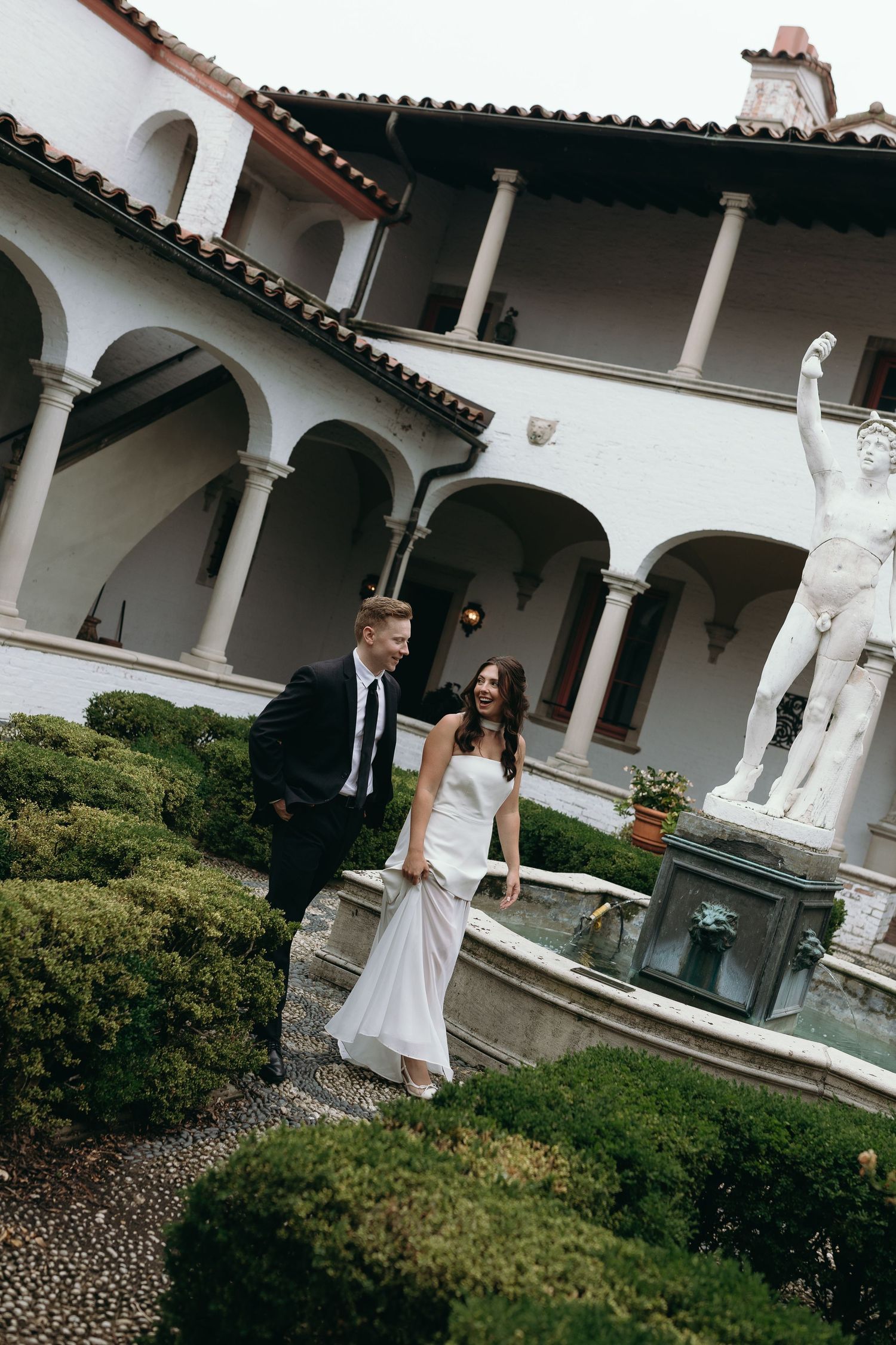 A wedding couple walks through a courtyard with white columns and statues at a Spanish-style villa.