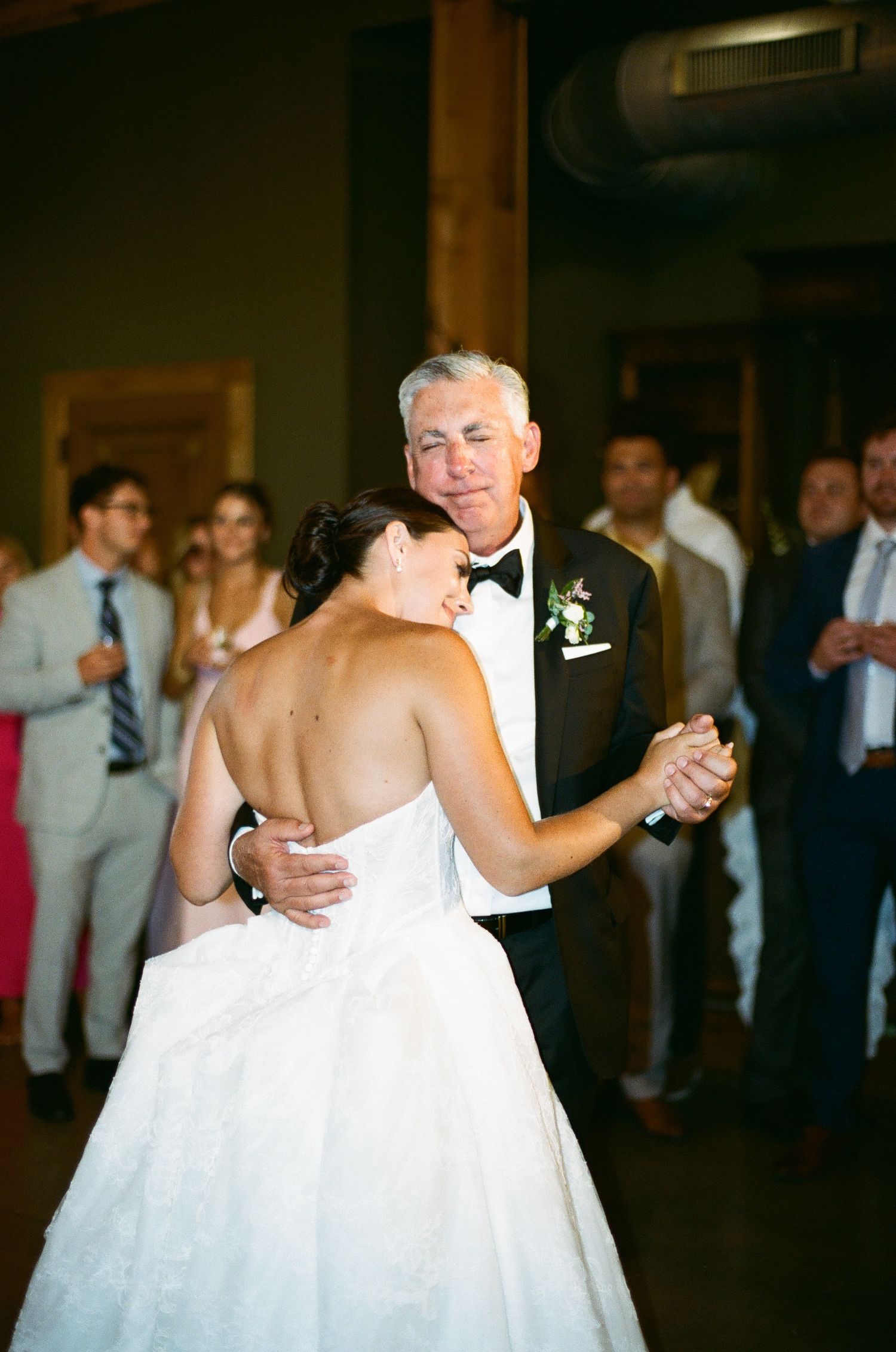 Bride and father share an emotional first dance at wedding reception, guests watching in background.