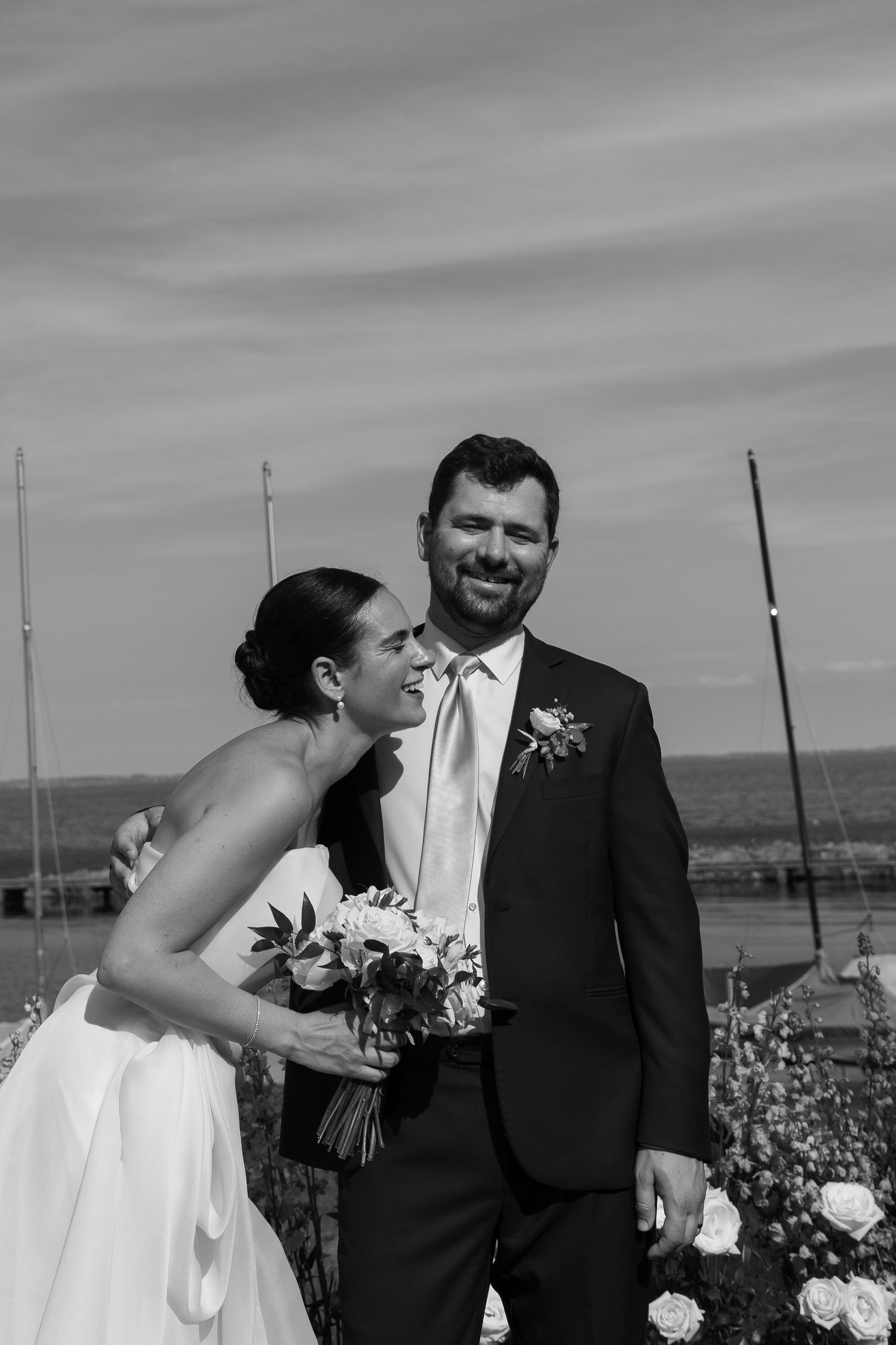 A black and white wedding portrait shows a happy couple outdoors sharing a candid moment by a waterfront.
