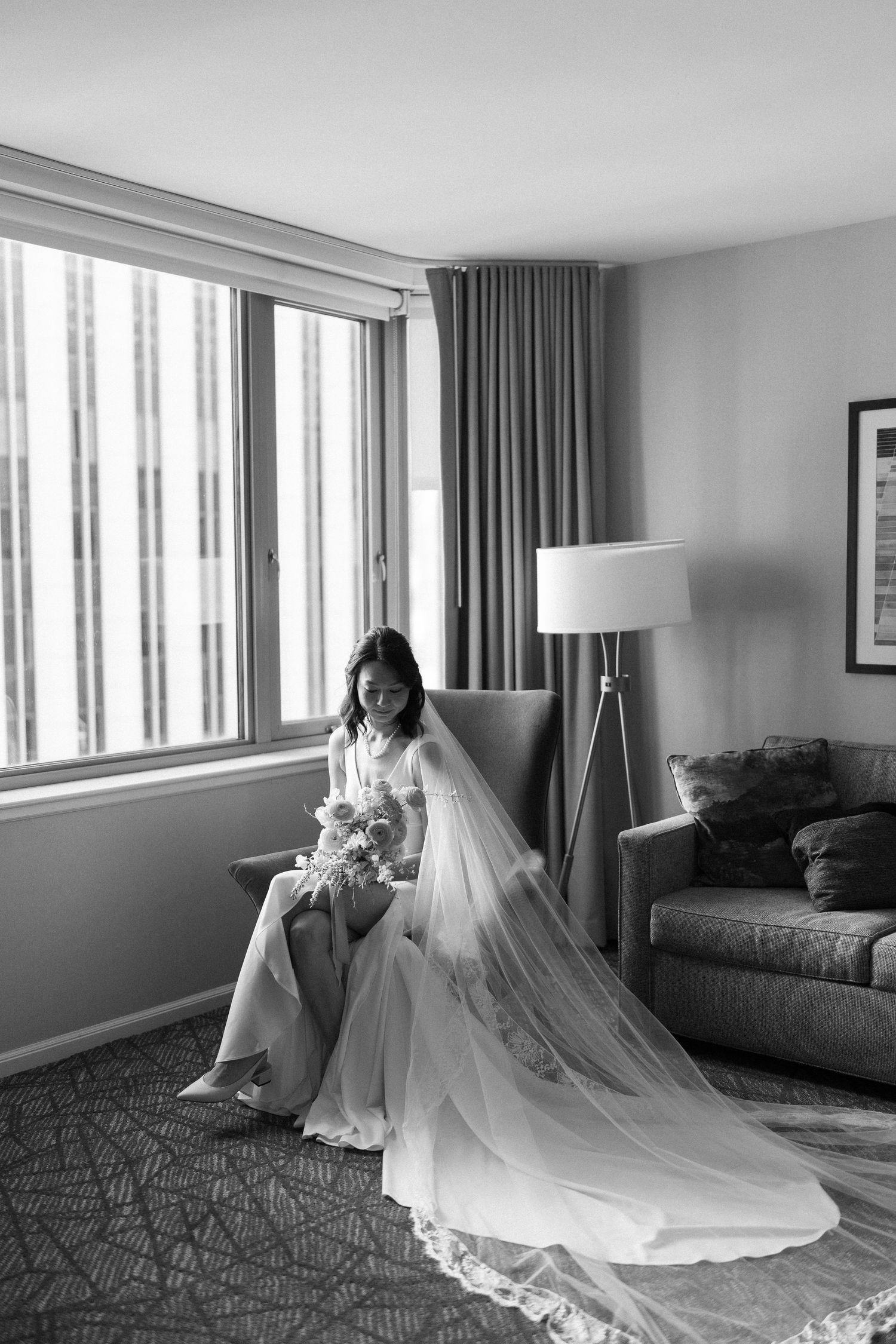 Black and white photo of bride in flowing wedding dress seated in hotel room.