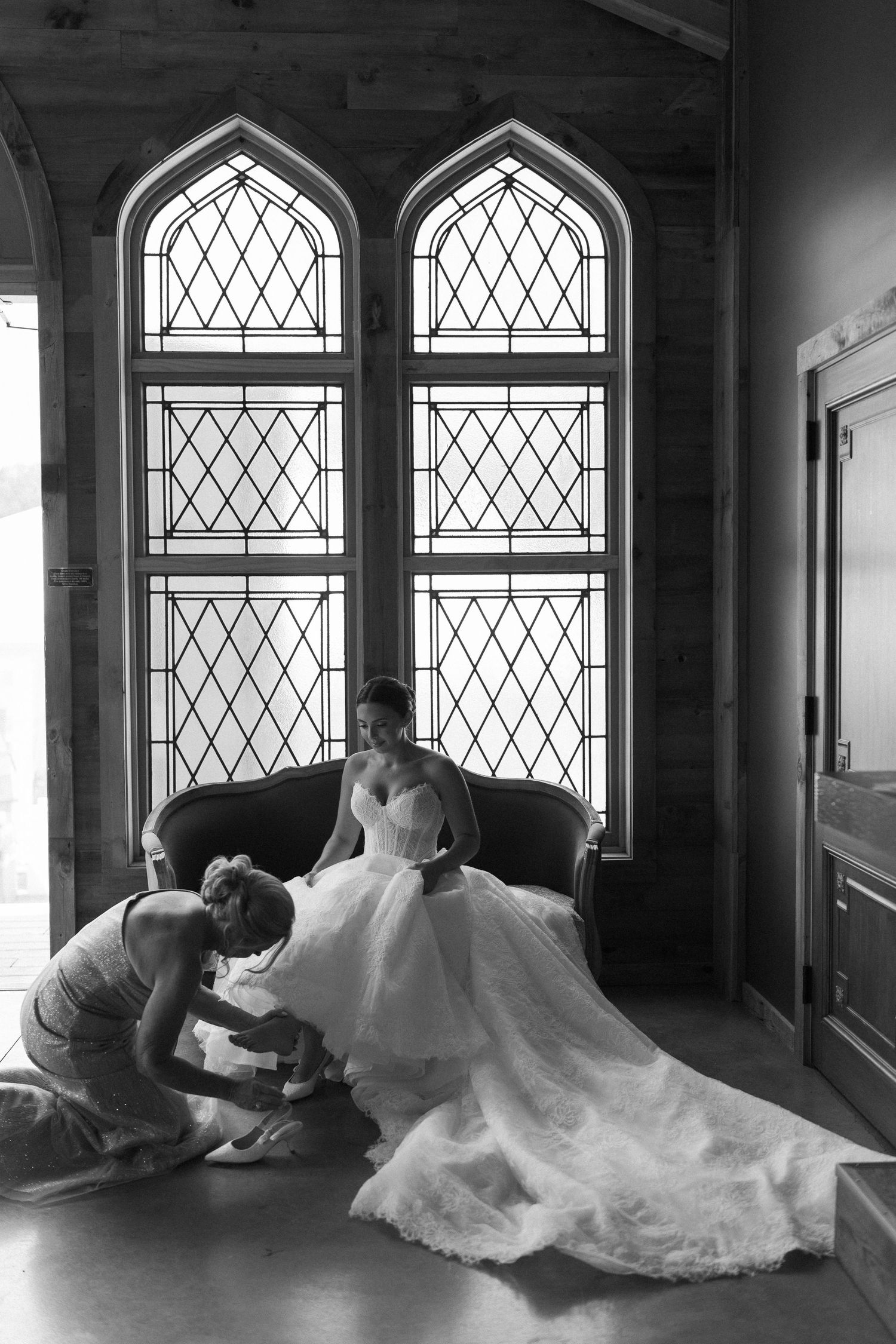 Bride in elegant white gown sits near gothic stained glass windows as bridesmaid adjusts her dress.