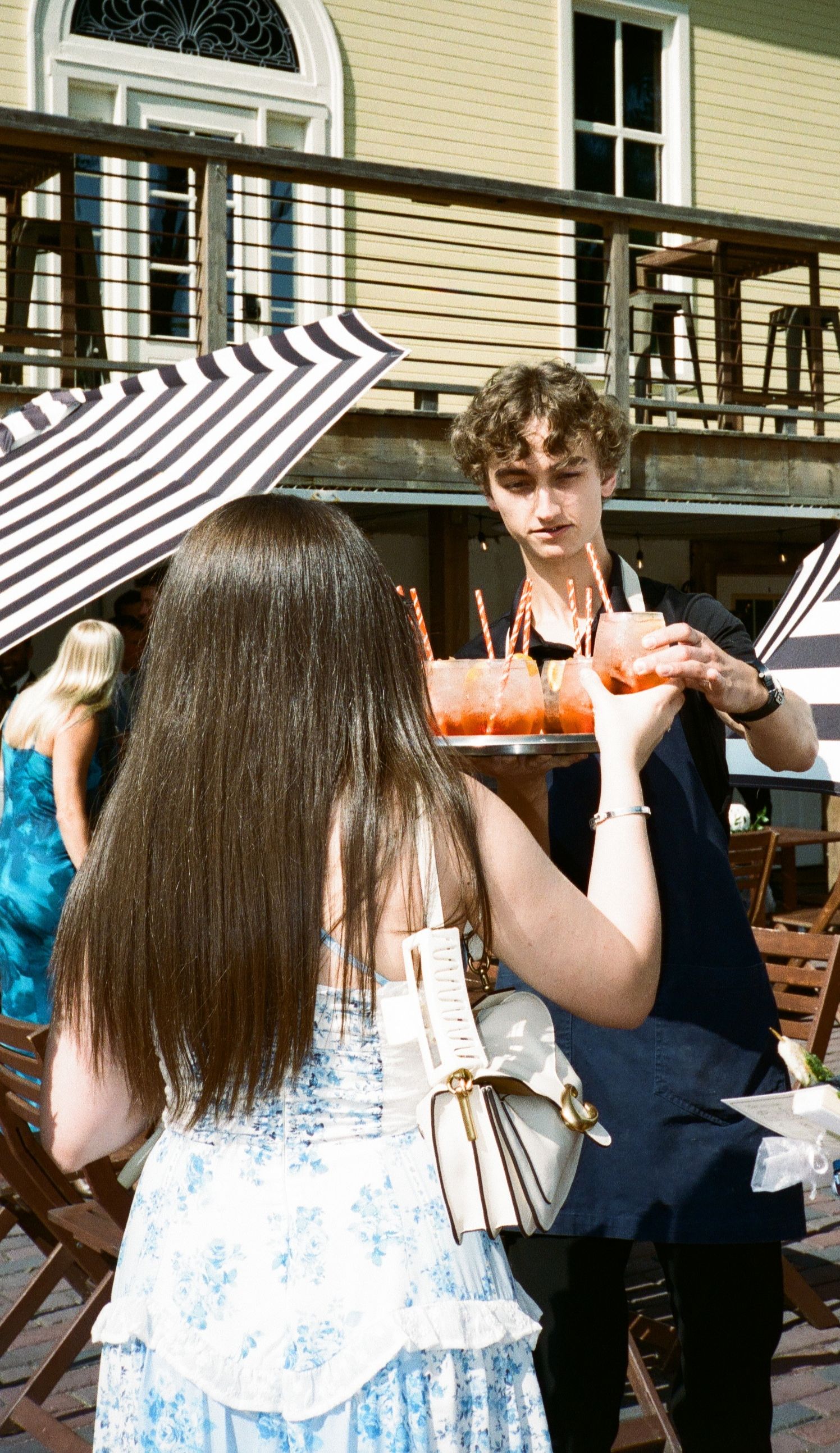 A young man serves colorful drinks on a sunny outdoor patio while a woman in a floral dress holds her purse.