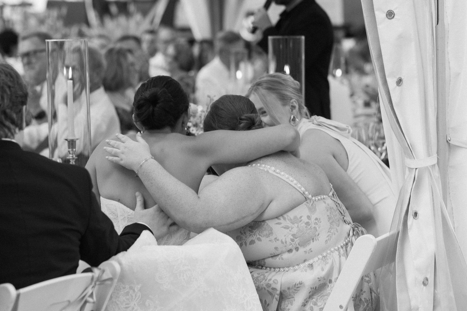 Black and white photo of three women in formal dresses sharing a warm embrace at a wedding reception.