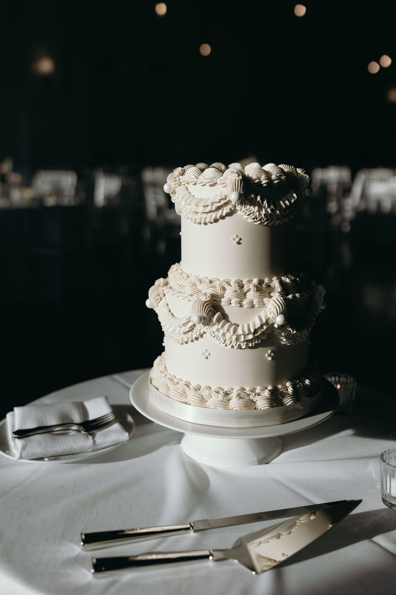 Three elegant white wedding cakes with pearl decorations displayed on tables with silver serving utensils.