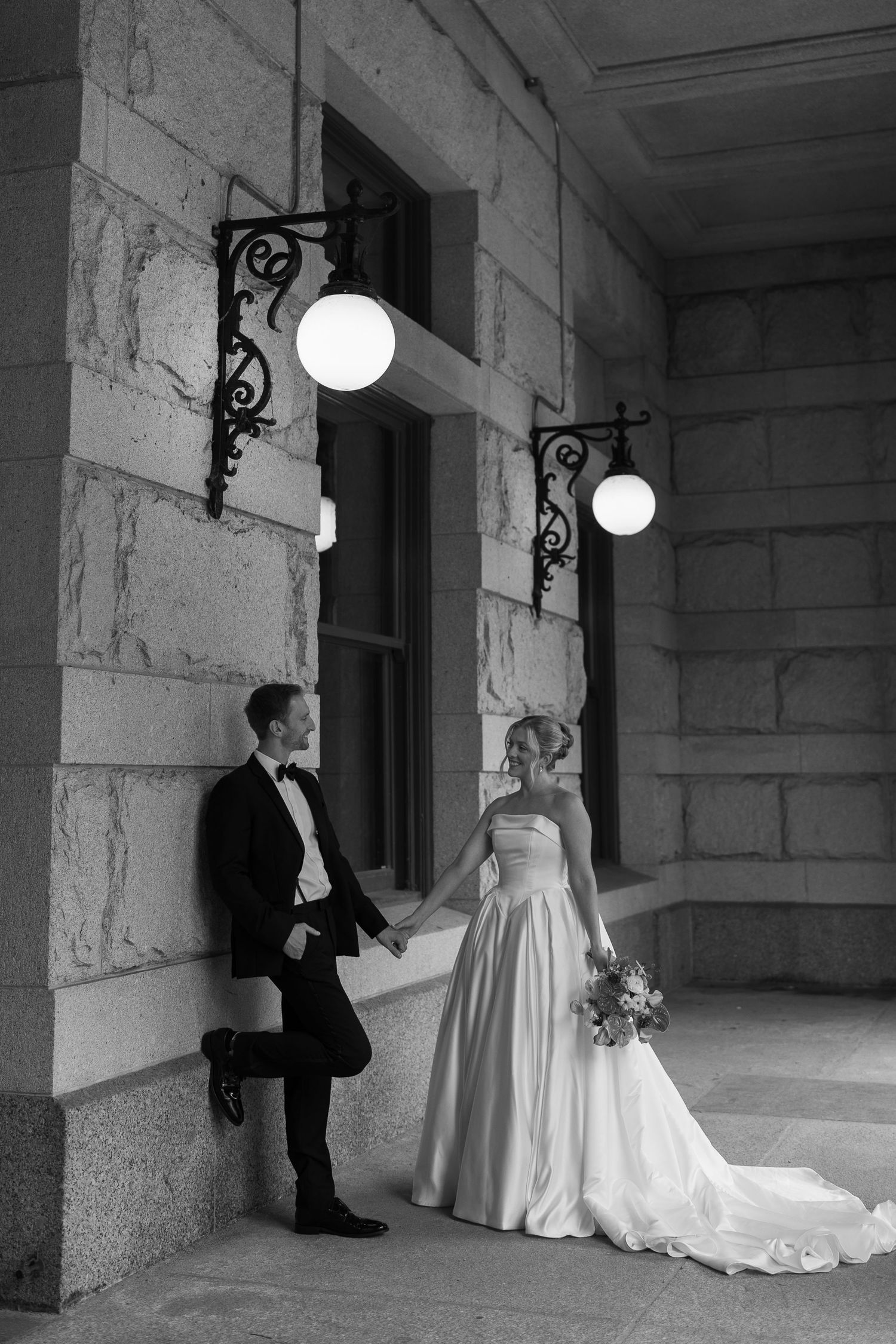 Elegant black and white wedding photo with couple standing beneath vintage light fixtures on stone building exterior.