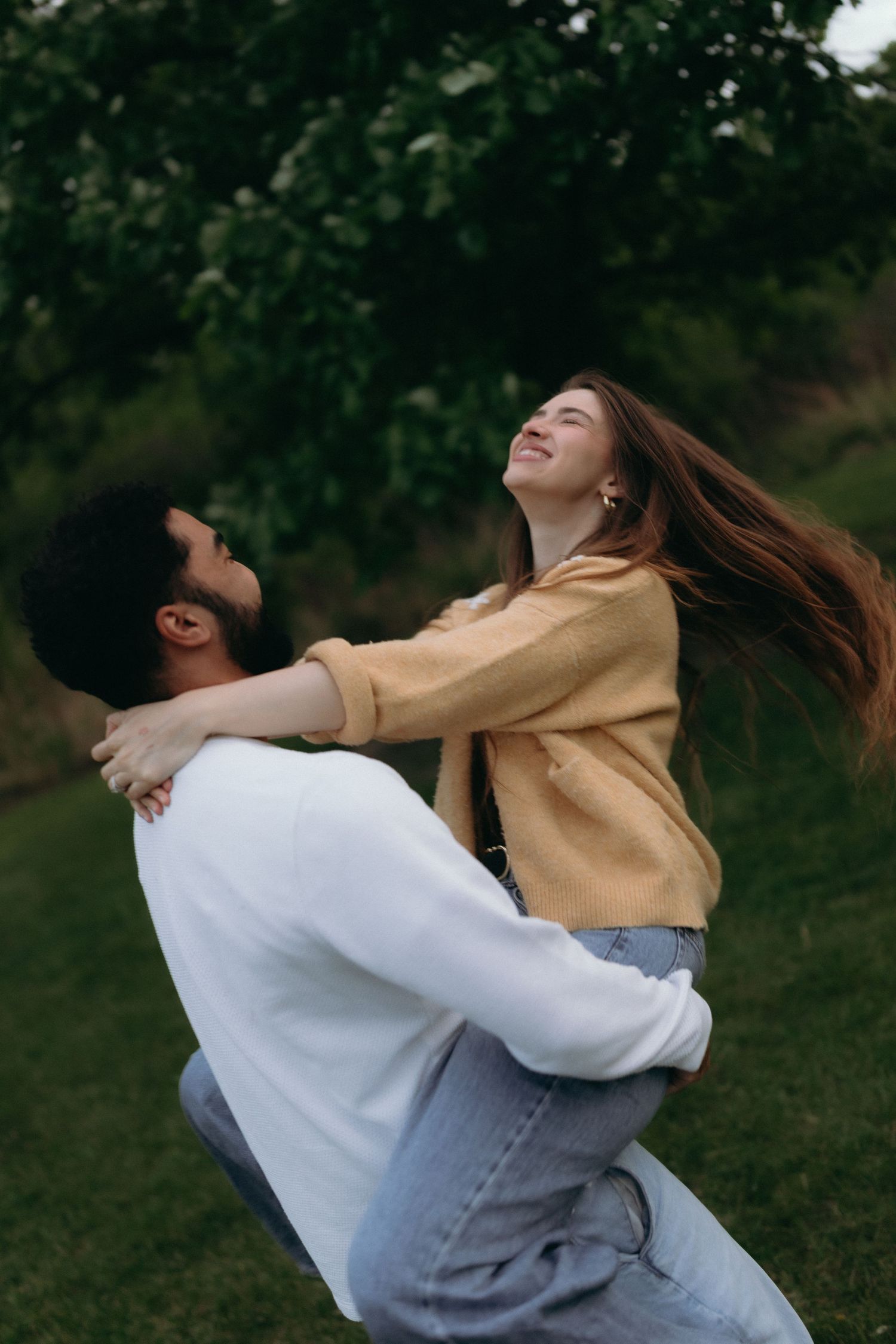 Chicago documentary engagement. A joyful couple dancing outdoors, man lifting woman in yellow jacket, both laughing in a lush green garden.