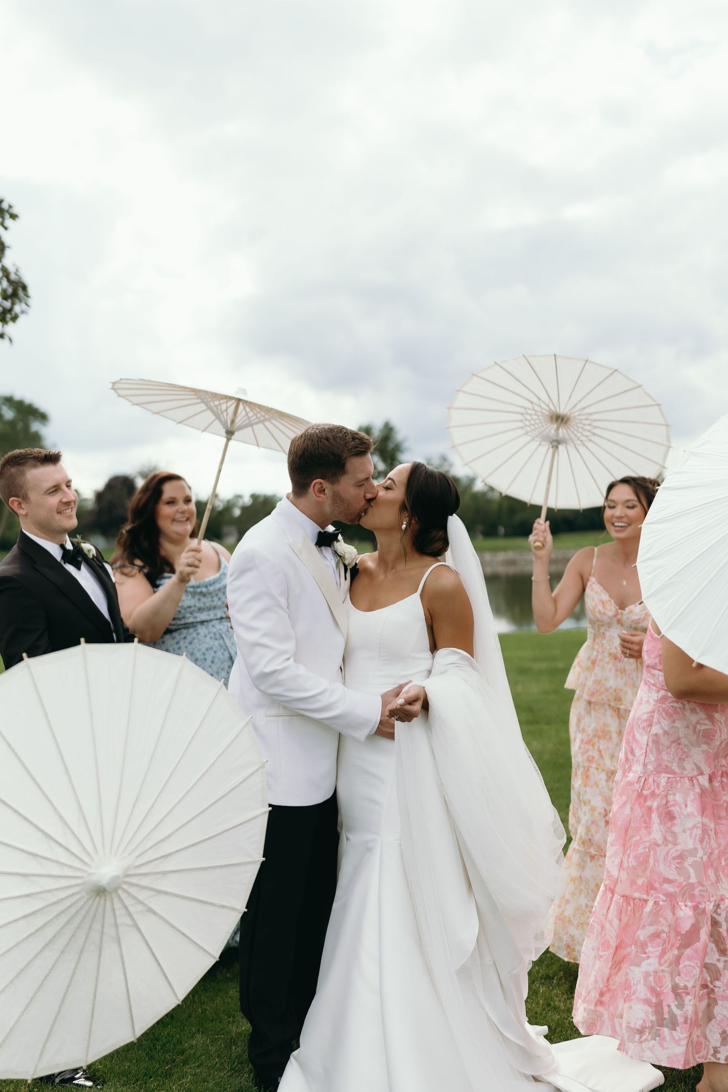 Wedding party poses with white parasols outdoors on cloudy day.
