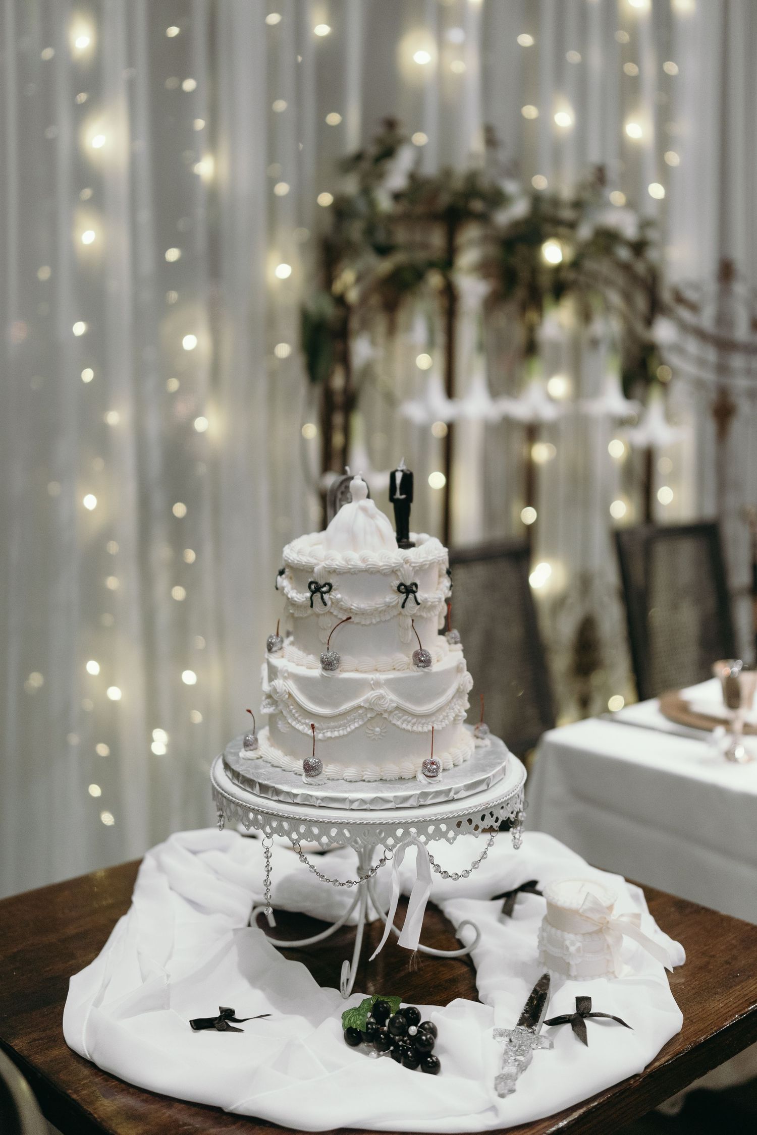 Classic white wedding cake with silver accents displayed against a backdrop of twinkling fairy lights.