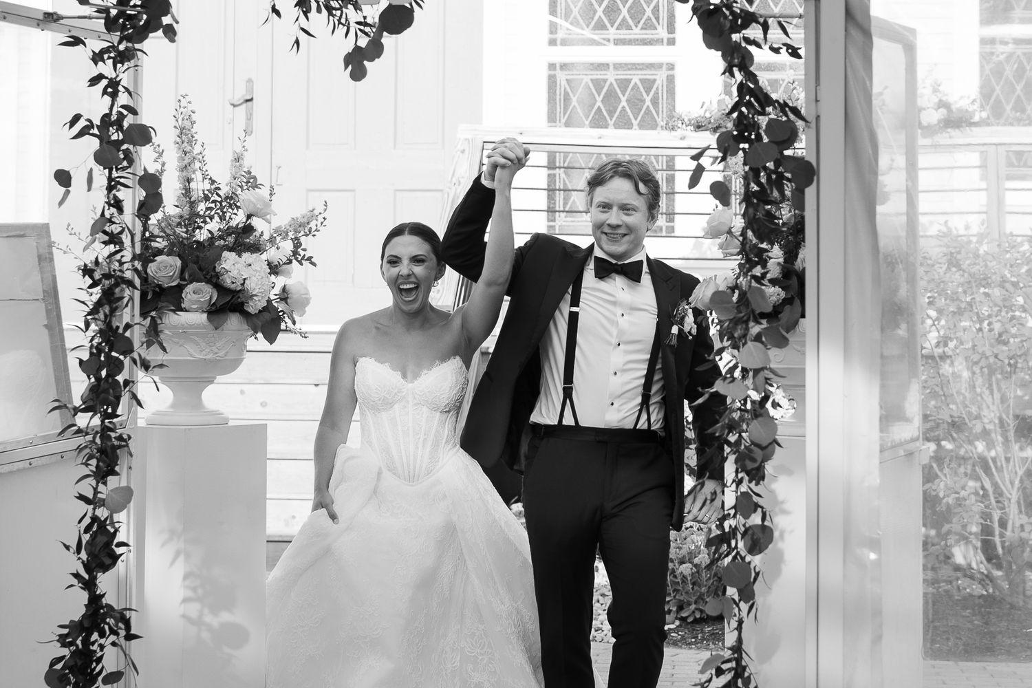 Black and white photo of a bride and groom celebrating under a floral arch at their elegant wedding ceremony.