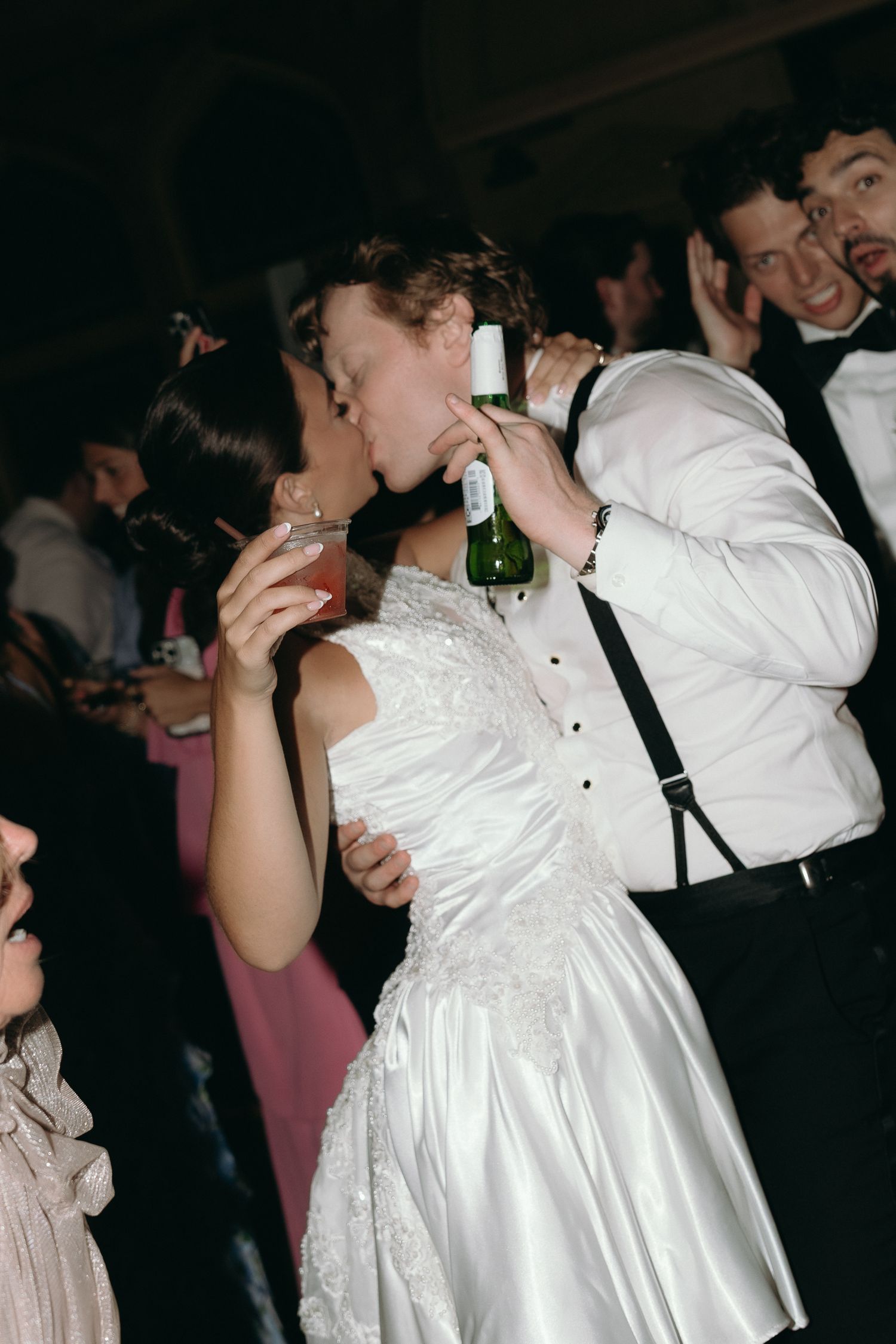 Bride and groom kissing while drinking champagne on wedding dance floor surrounded by guests.