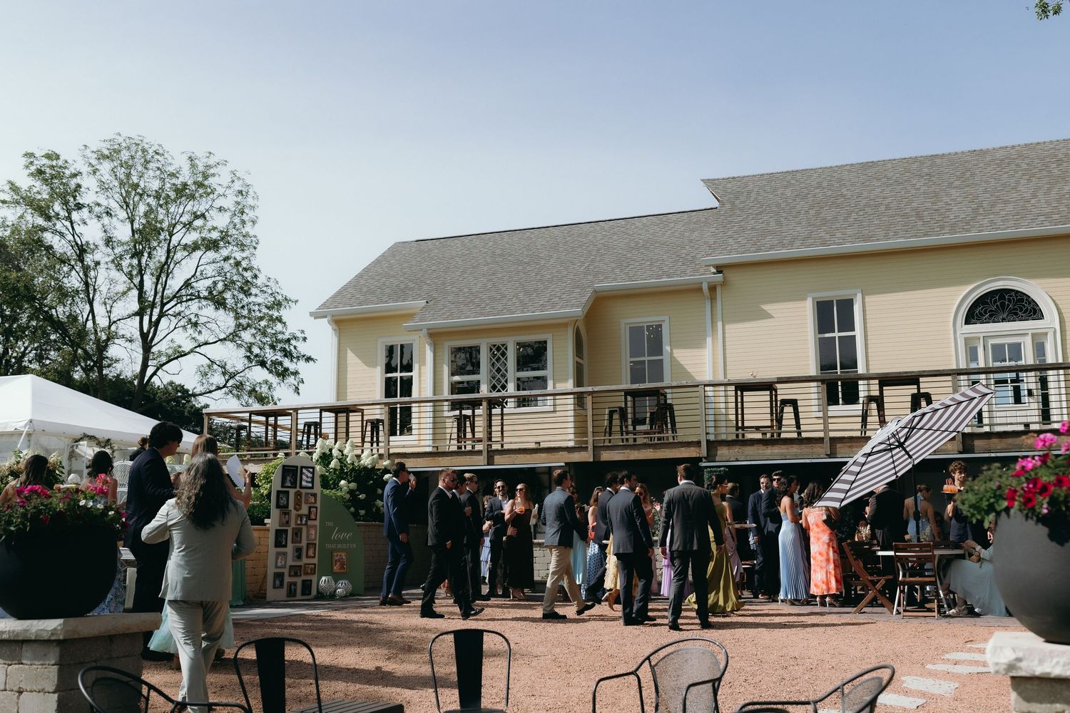 Guests mingle outdoors at a yellow venue with a deck during a wedding reception on a clear day.