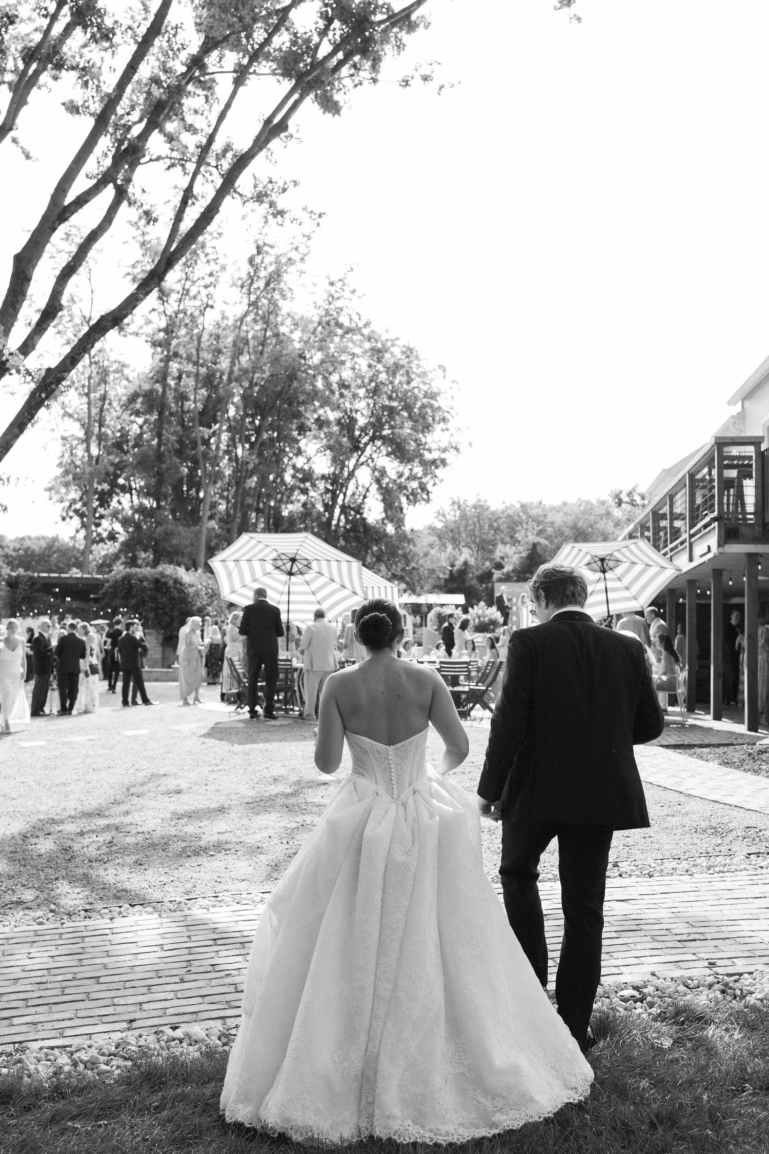 Black and white photo of bride and groom walking hand in hand toward outdoor wedding reception with umbrellas and guests.