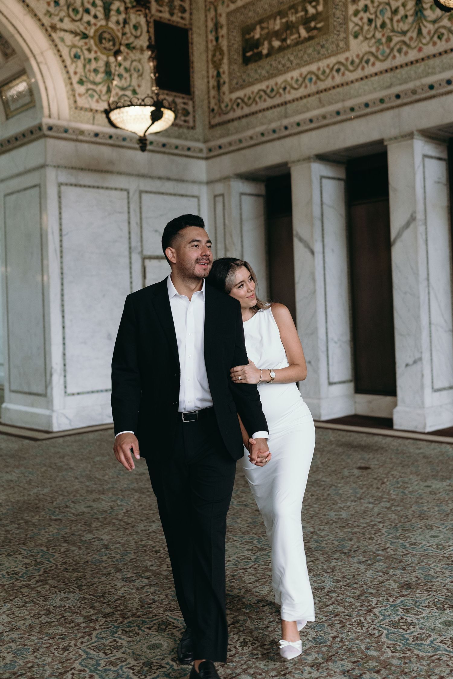 Couple in elegant black and white formal attire walking together through an ornate columned hallway with decorative ceiling.
