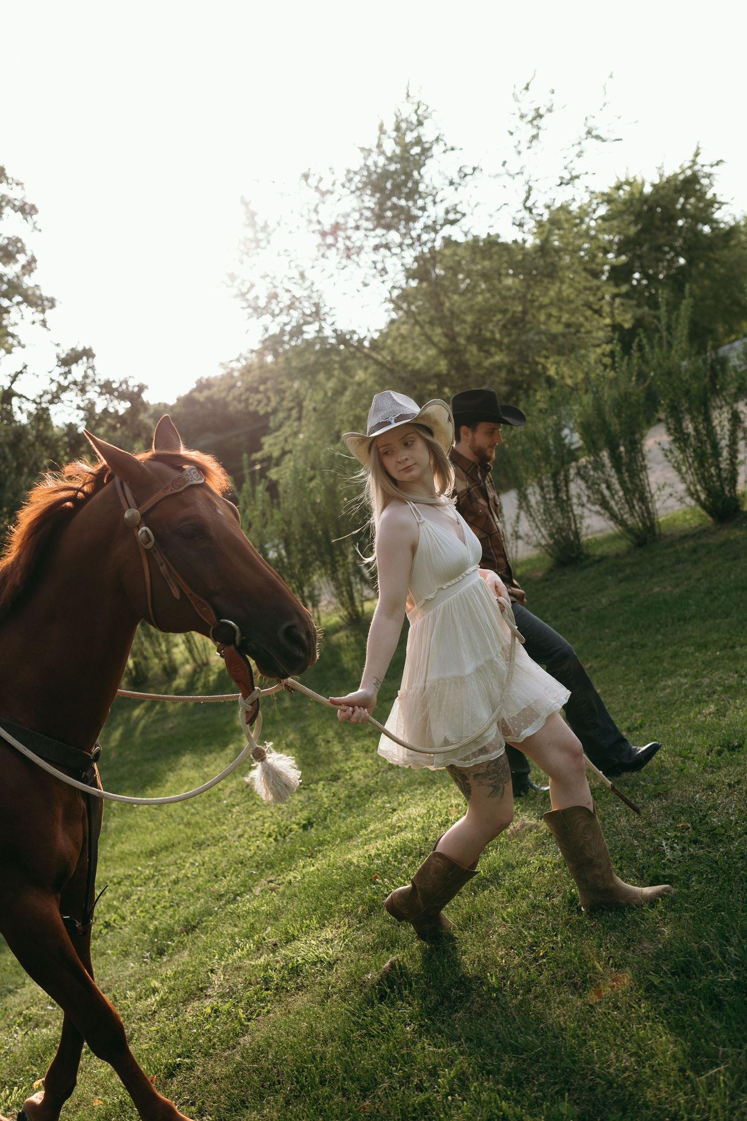 A couple in western attire walks with their horse on a grassy hill during golden hour lighting.