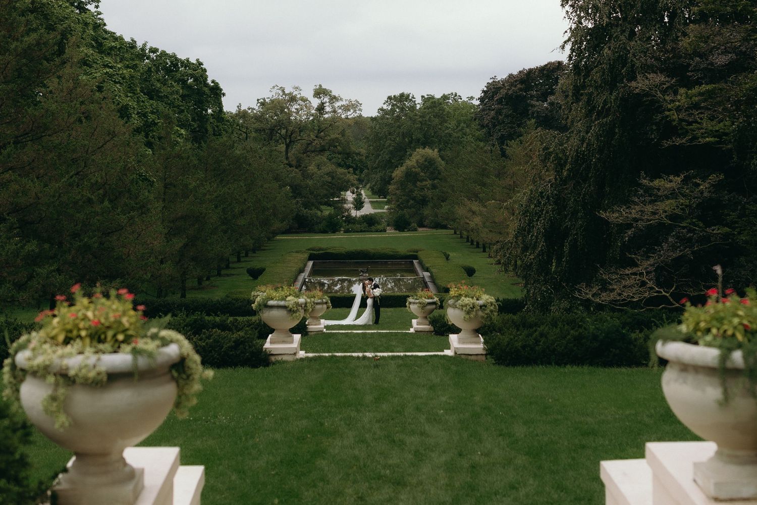 Elegant formal garden with manicured hedges, ornate stone urns, and a couple standing in the distance on a lush green lawn.