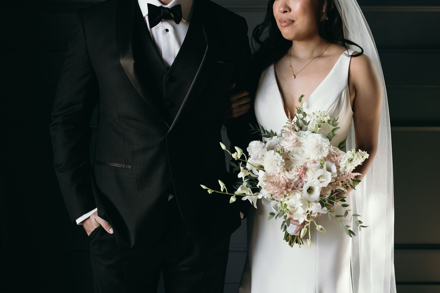 Elegant wedding couple in black tuxedo and white gown standing together with blush and white floral bouquet.