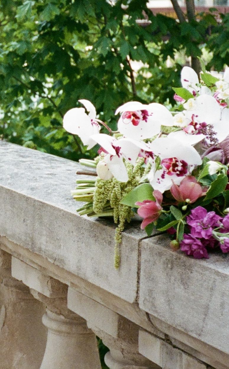Wedding bouquet with white orchids and purple flowers resting on a stone balustrade with green trees in background.