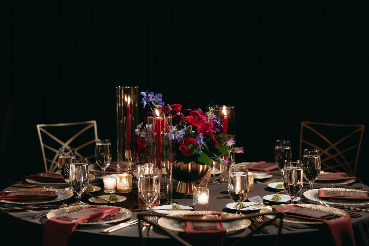 Elegant dark wedding reception table with red floral centerpiece, gold candles, crystal glasses, and crimson napkins.