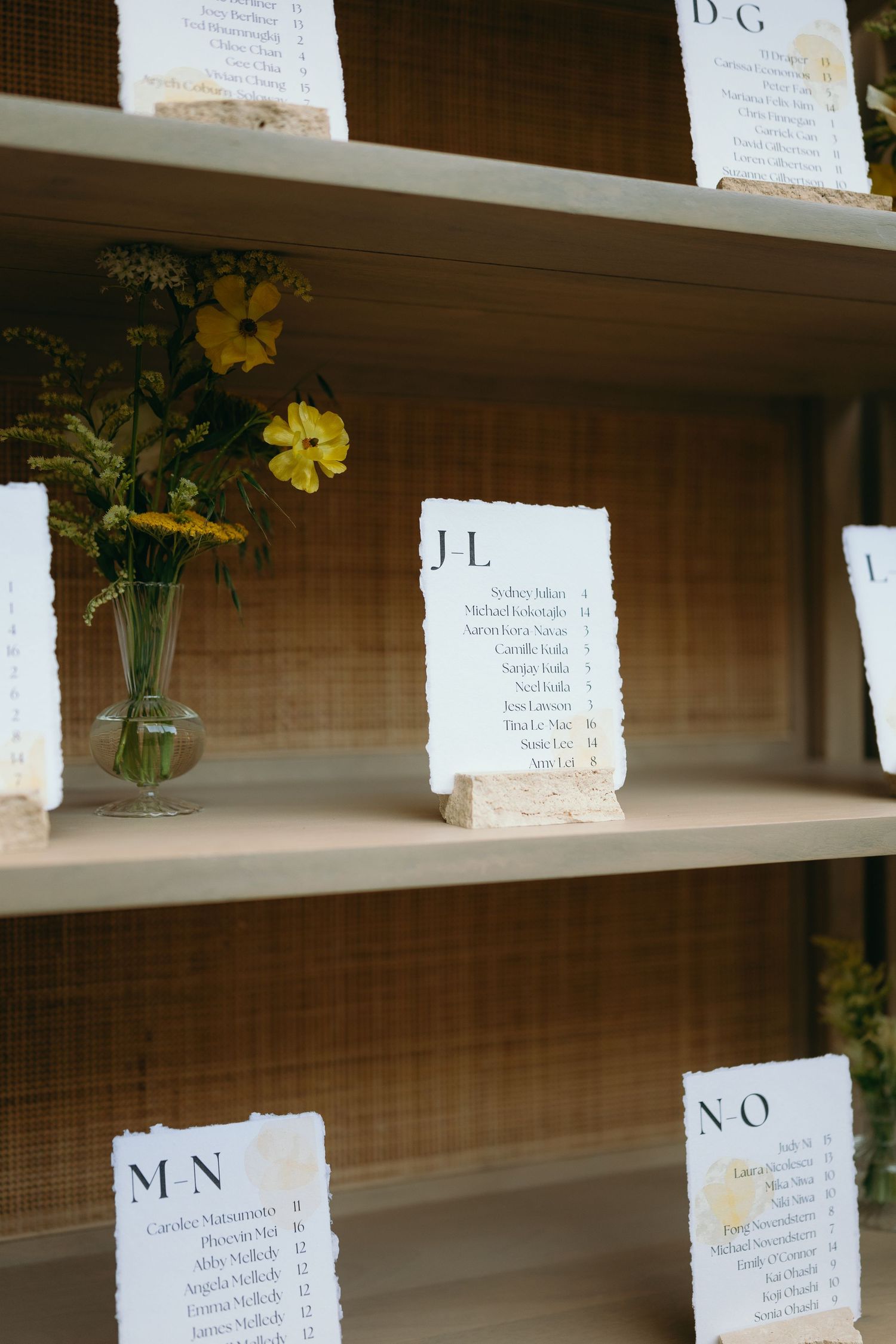 Wedding seating chart display with yellow flowers and alphabetized name cards on wooden shelves.