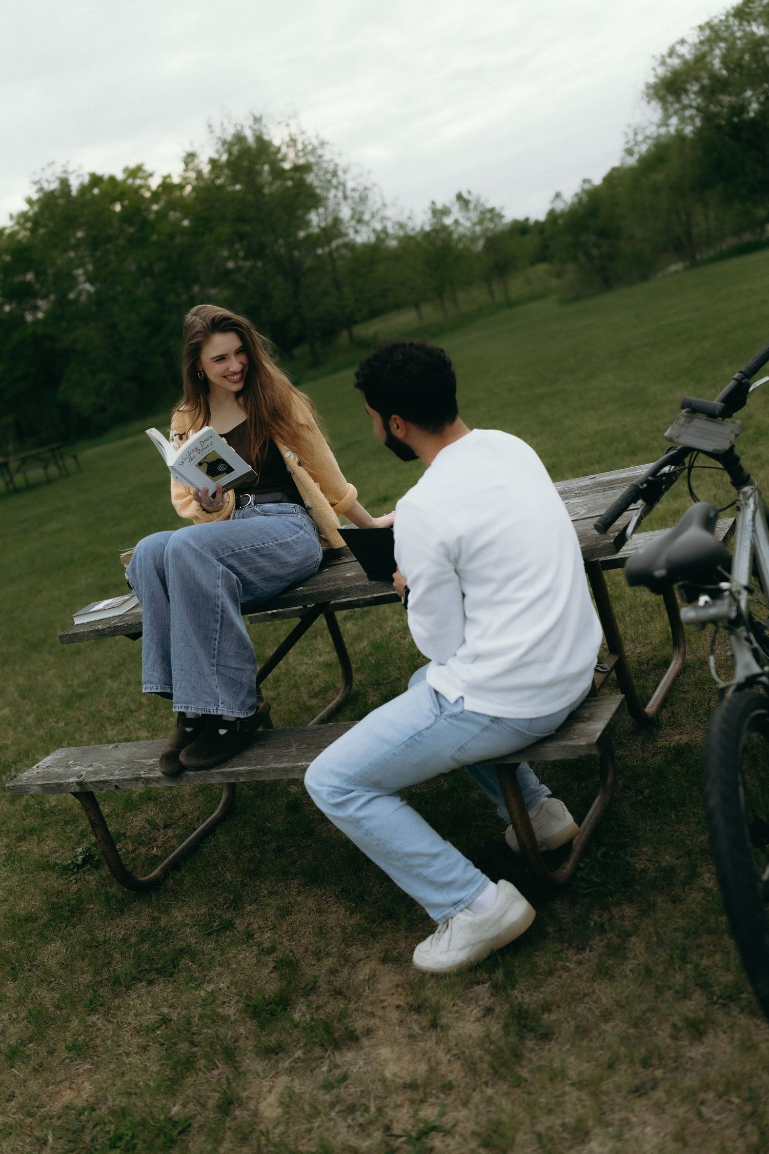 A couple sits at a wooden picnic table in a park, with a woman reading a book and a bicycle nearby.
