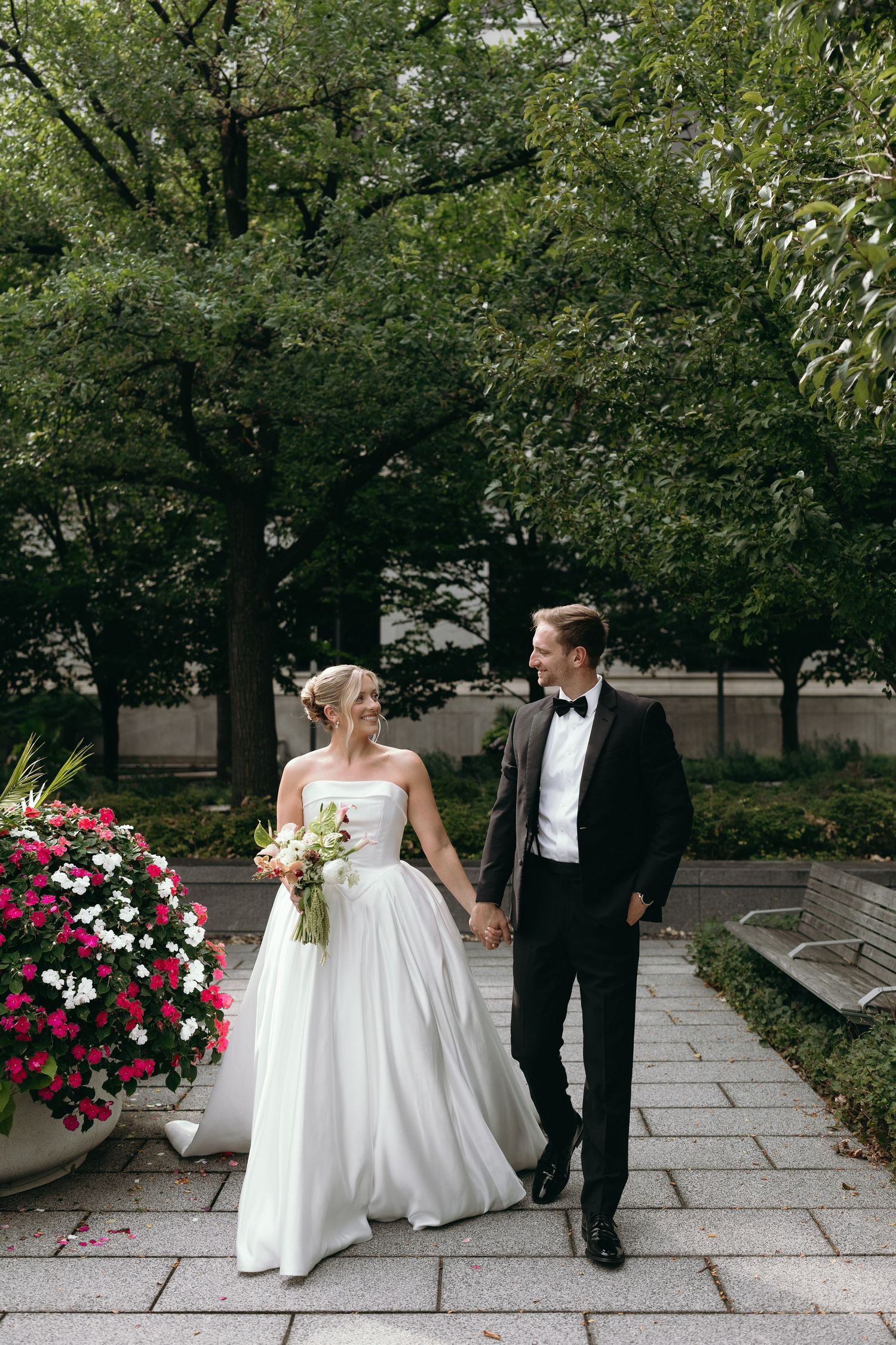 Newlyweds walking hand in hand on stone path with lush greenery and pink floral arrangements in background.