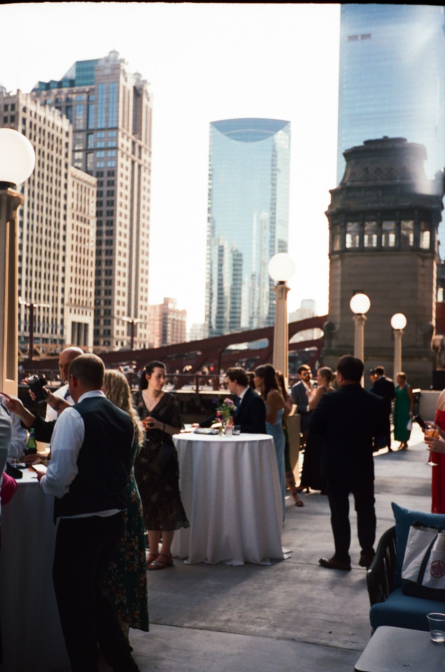 Elegant rooftop cocktail hour with city skyline backdrop, guests mingling around white draped tables in urban setting.